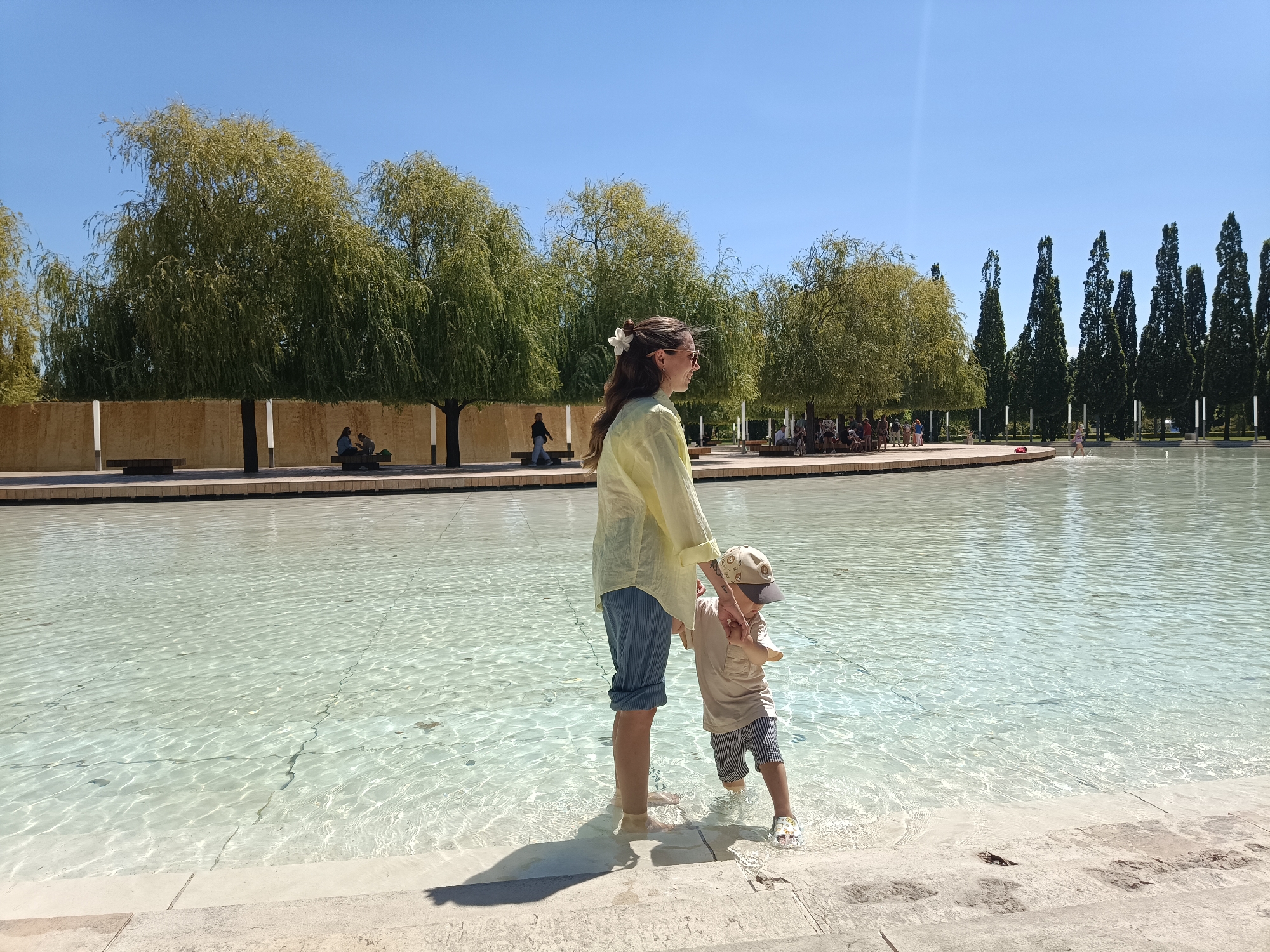 The image depicts a clear, sunny day with a bright blue sky, showcasing a shallow pool of water with visible ripples and sunlight reflecting off its surface. A woman wearing a light yellow shirt and blue striped pants stands in the water, holding the hand of a young child. The child, dressed in a light-colored t-shirt, striped shorts, and a baseball cap, is also standing in the water. Both are facing to the right of the frame. In the background, there are trees, a wall, and several people are seated on benches or walking. Further in the background, a line of tall, dark green trees stands against the horizon. The scene is an outdoor public space, likely a park or recreational area. There is no visual information in the image that suggests the location is Krasnodar, Russia.