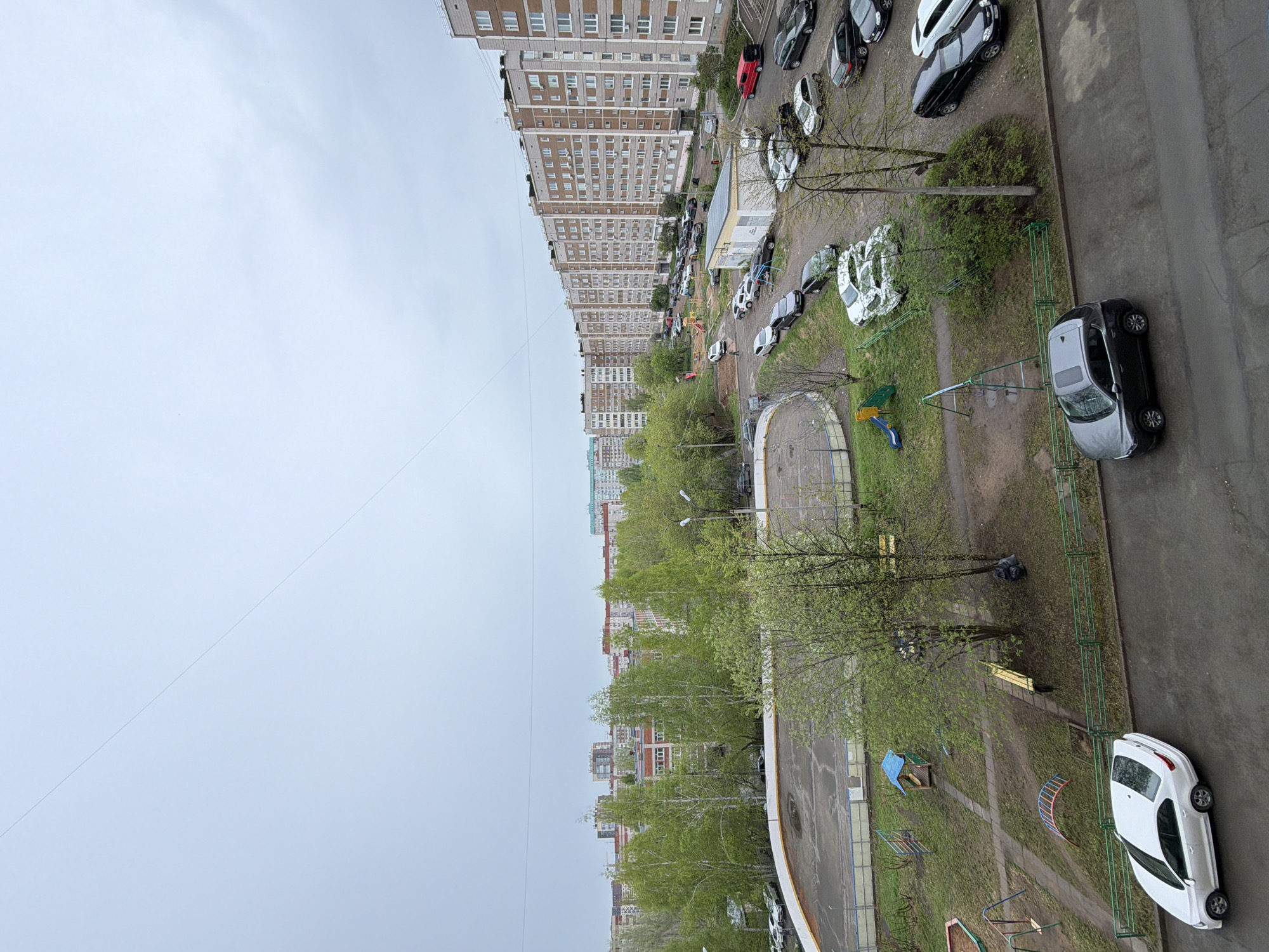 The image shows an elevated view of an urban landscape in Izhevsk, Russia. A row of multi-story residential buildings lines the left side of the image, extending into the distance. A street runs along the right side, occupied by parked cars. Between the buildings and the street is a green space containing trees, grass, and playground equipment. The sky is overcast.