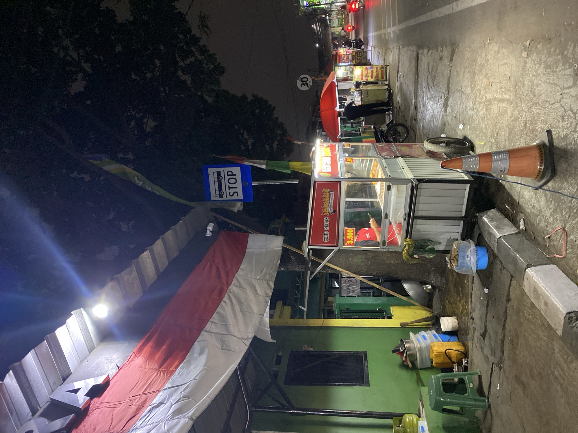 The image depicts a street scene at night in Tangerang, Indonesia, likely a commercial area with food vendors. A prominent Indonesian flag hangs from a building on the left. In front of the building, there are several food carts. One cart, labeled "GEHU PEDAS" and displaying a price of "3.500," is in the foreground. Another cart with a red umbrella is also visible, with a person attending to it. Further down the street, more food carts and a speed limit sign indicating "30" are present. A bus stop sign with the word "STOP" is also visible. The street is paved, and there is a sidewalk with a concrete curb. A traffic cone is placed on the sidewalk near the food carts. The overall atmosphere is dimly lit, suggesting nighttime.