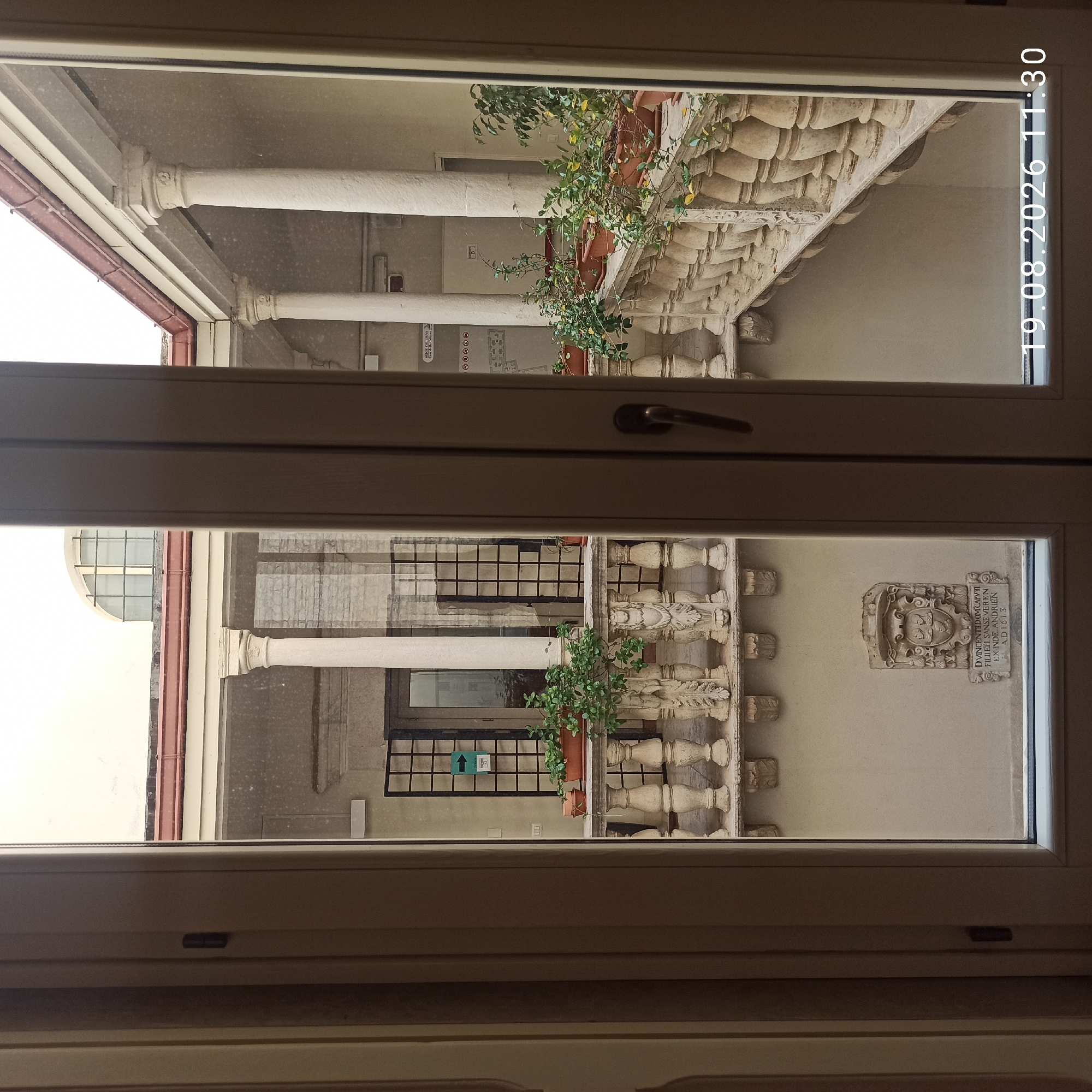 The image shows a view through an open doorway or window into an interior courtyard of an old building. The courtyard features a covered walkway with ornate stone columns and a decorative stone balustrade running along the upper level. Potted plants are placed on the balustrade. On the wall of the courtyard, there is a stone carving with text, and an arrow sign pointing left. The architectural style suggests a historical building, potentially a museum or civic building. The timestamp in the lower right corner indicates the image was taken on August 19, 2026, at 11:30. Based on the architectural style and the presence of historical inscriptions, this scene is likely located in the historic center of Ruvo di Puglia, Italy, possibly within the Palazzo Caputi, which houses the Museo del Libro (Book Museum) and features similar architectural elements and historical artifacts. There are no people visible in the image.