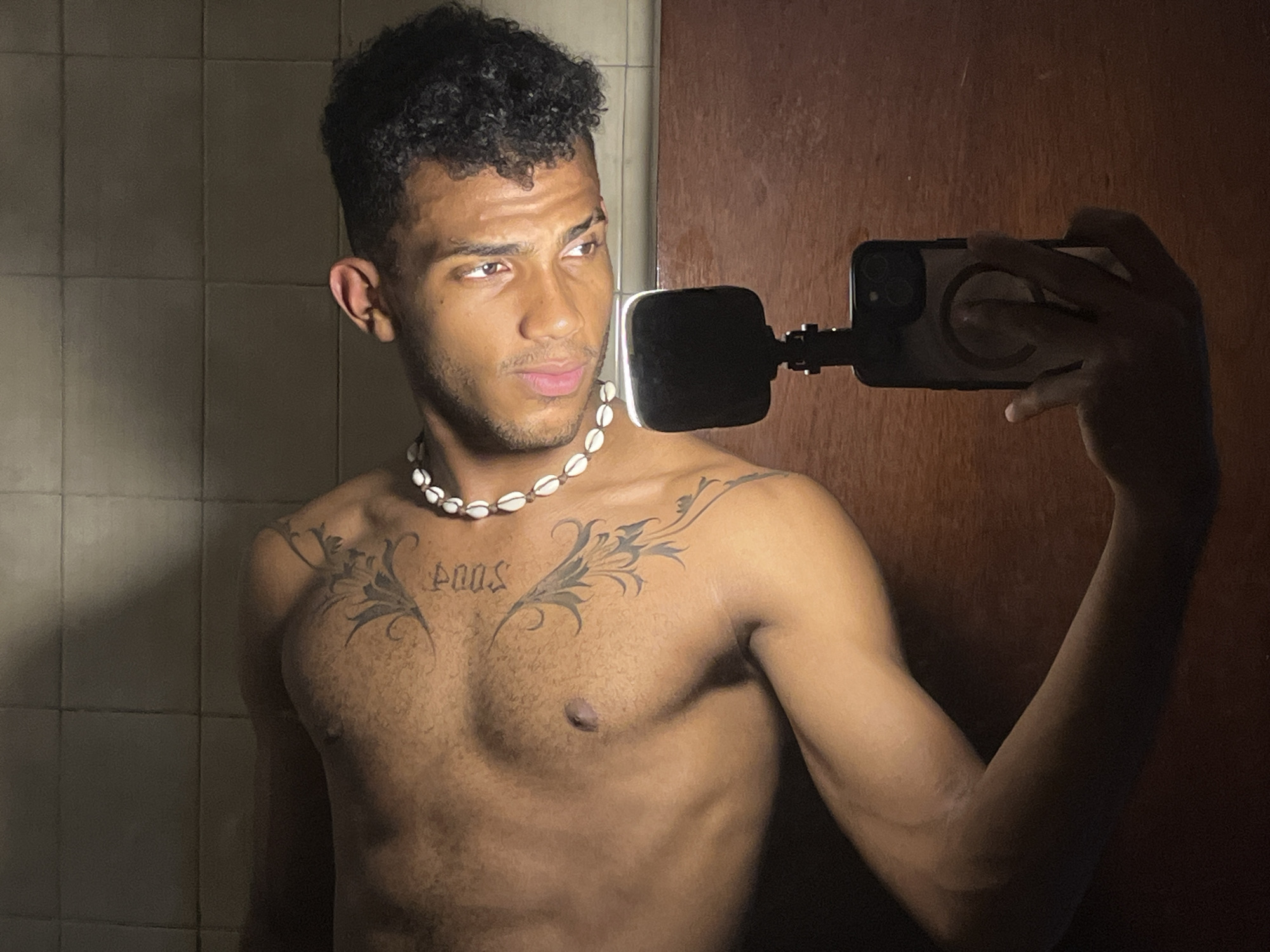 A dark-skinned man with curly hair is taking a selfie. He is shirtless and wearing a necklace made of shells. He has a tattoo on his chest. He is holding a smartphone with a black case. The background includes a tiled wall and a brown door. The location is likely indoors, but the exact location within Porlamar, Venezuela, cannot be determined from the image.