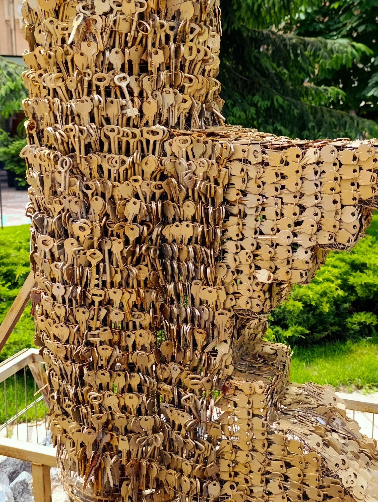 The image shows a sculpture made of numerous keys, located outdoors in Kyiv, Ukraine. The sculpture is constructed from many small, brass-colored keys, attached to a wire frame. The keys are arranged in a dense, layered fashion, creating a textured surface. The sculpture appears to be abstract in form, possibly resembling a stylized animal or object due to its rounded shape and projecting elements.
The background consists of lush green foliage, including bushes and trees, suggesting a park or garden setting. Sunlight illuminates the sculpture, casting subtle shadows and highlighting the metallic sheen of the keys. In the foreground, a railing and some rocks are visible, further indicating an outdoor public space. The overall impression is that of an artistic installation in a natural environment within the city.