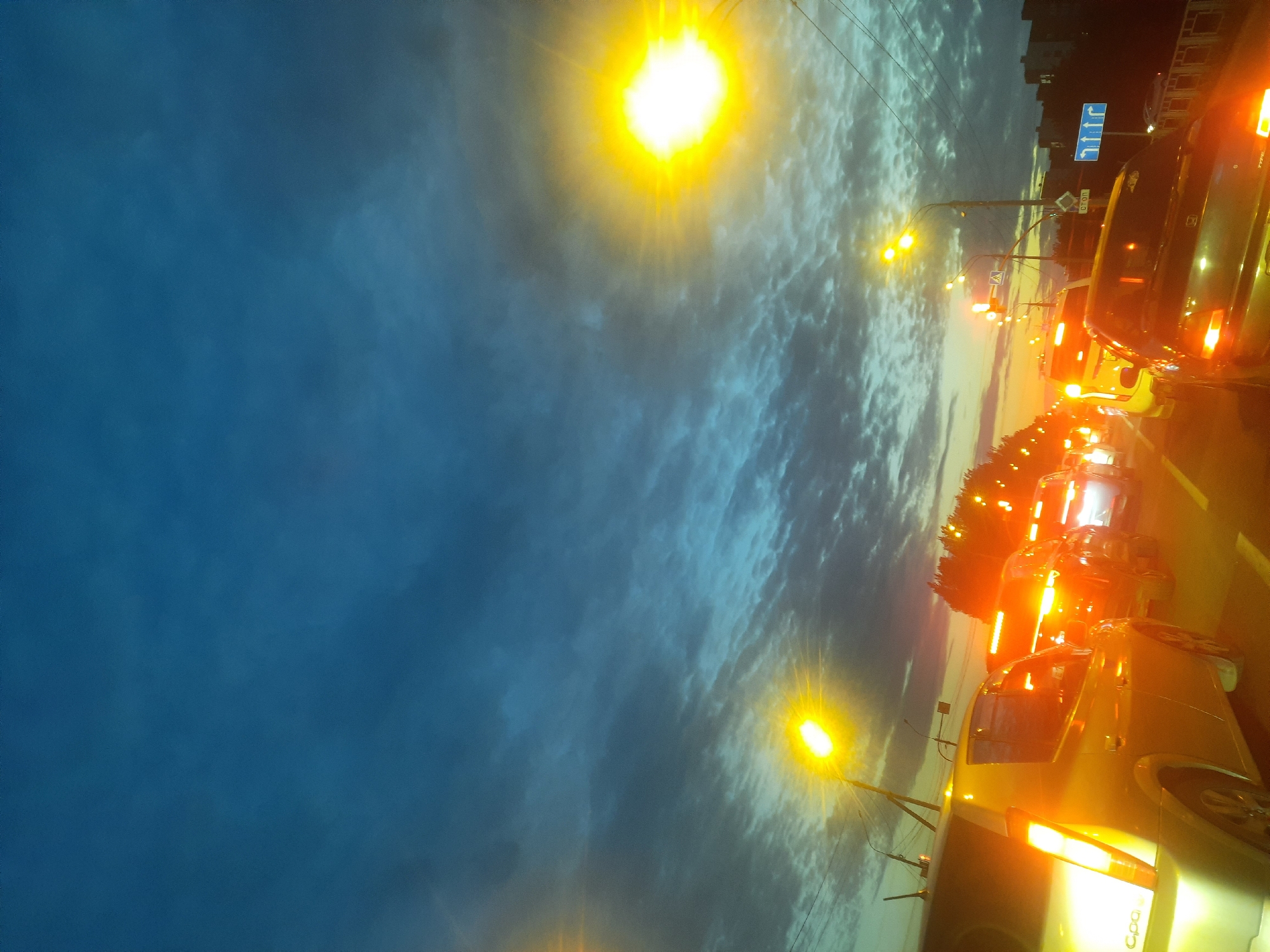 A nighttime traffic jam is captured on a bridge in Barnaul, Russia. The sky is a deep blue with streaks of clouds, illuminated by streetlights and the taillights of cars. Several cars are visible, including a silver sedan in the foreground with its taillights on. The road appears to be a multi-lane highway, and the bridge railing is visible on the right side of the image. Streetlights line the road, casting a warm glow on the scene. The overall impression is one of urban bustle at dusk.