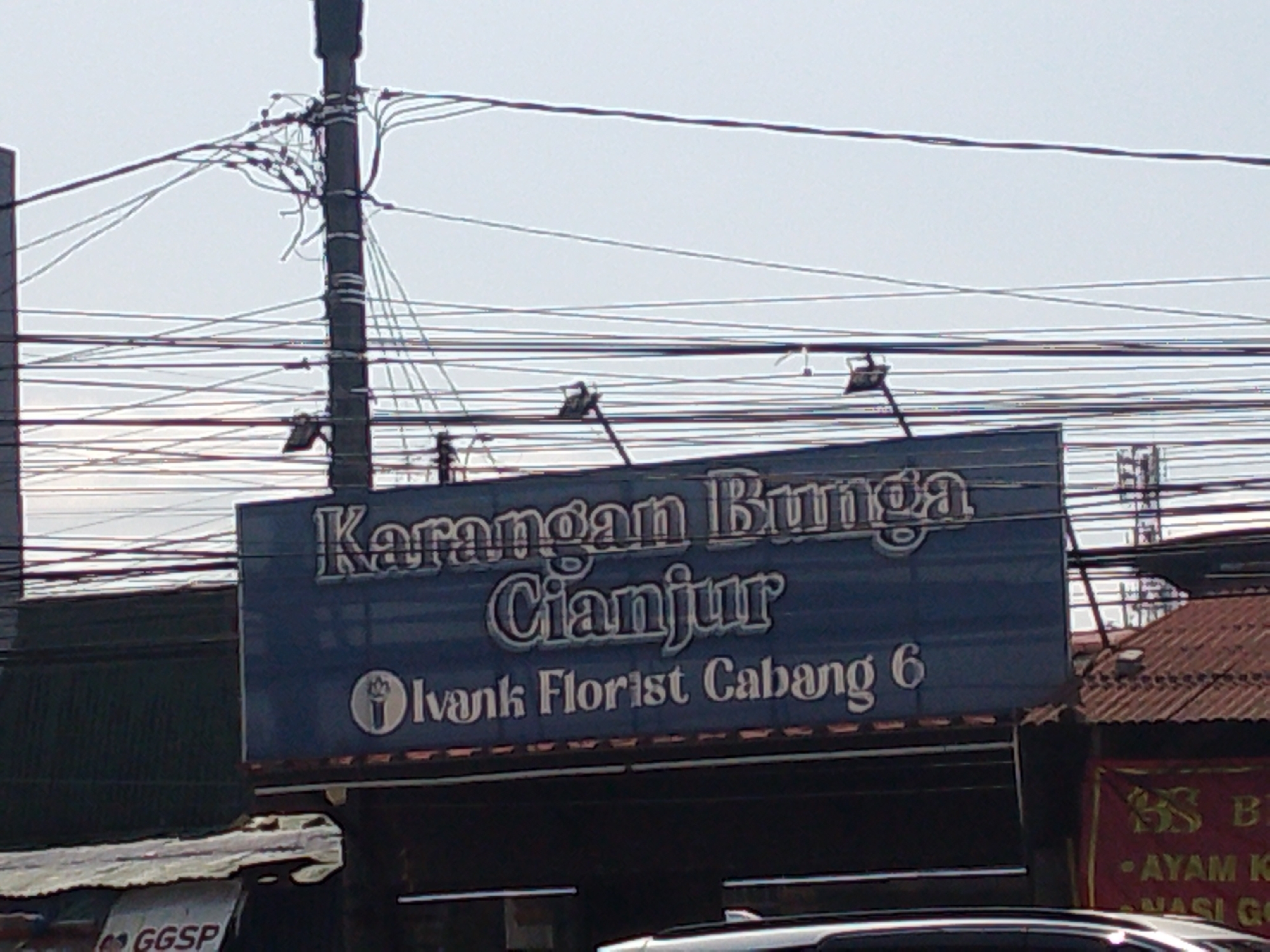The image shows a dark blue sign with white lettering for "Karangan Bunga Cianjur" and "Ivank Florist Cabang 6". The sign is mounted on a building with a corrugated metal roof. There are numerous electrical wires overhead, and a utility pole is visible to the left of the sign. To the right of the florist sign, a red banner with gold lettering is partially visible, displaying "88 B" and "- AYAM K". The overall environment appears to be an urban street scene in a developing area. The lighting suggests it is daytime.
