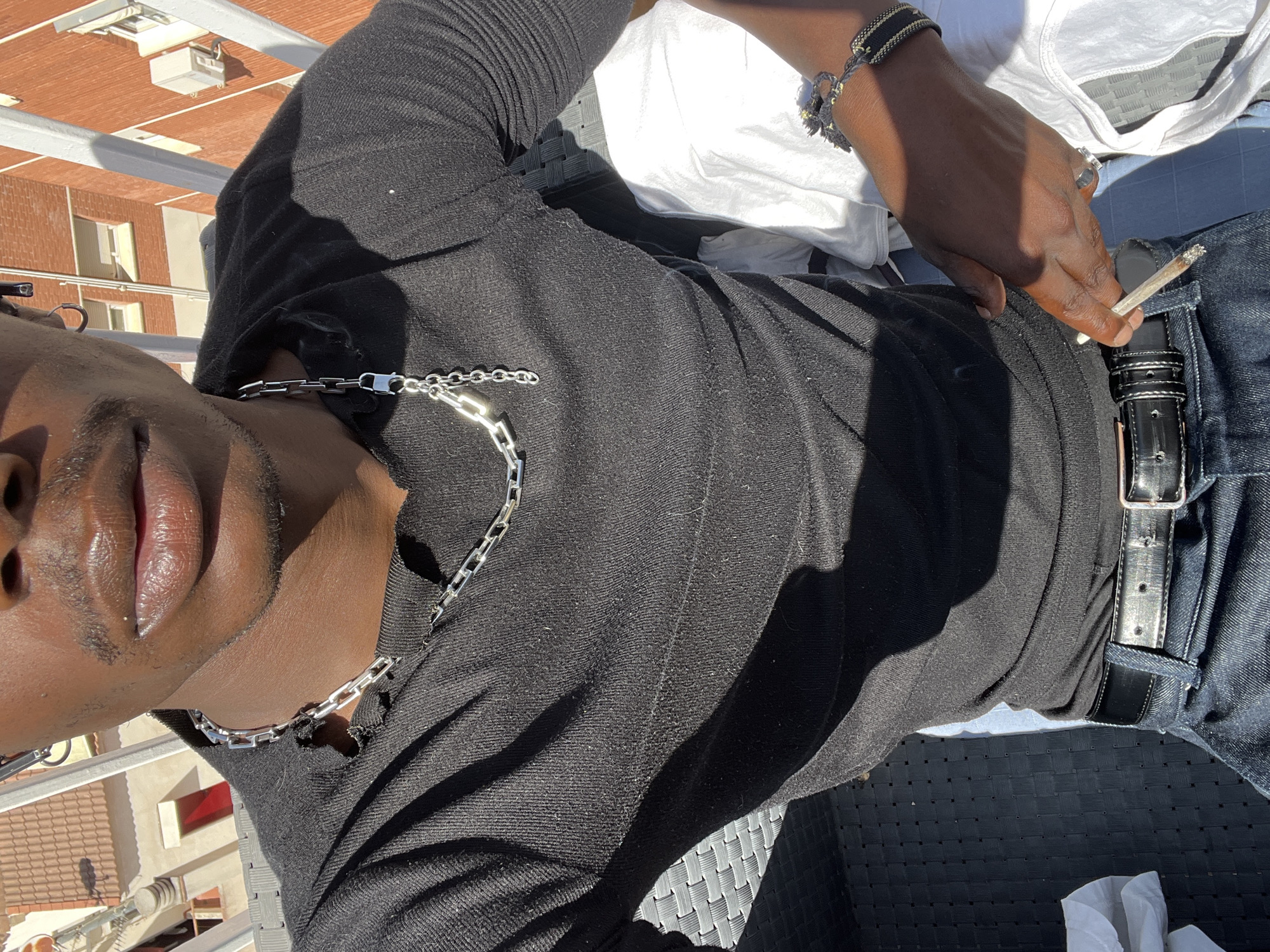 The image captures a person of color reclining outdoors, likely on a balcony or terrace, under bright sunlight. The shot is angled, presenting the subject diagonally across the frame.
The person is wearing a black, long-sleeved top with a visible texture, and dark blue denim jeans secured with a black leather belt and a silver buckle. Accessories include a thick, rectangular-linked silver chain around their neck, a silver ring on their right hand, and several bracelets (some beaded, some woven) on their right wrist. In their right hand, they hold a lit cigarette or hand-rolled joint, with a visible ash tip. A folded white piece of fabric, possibly a t-shirt or towel, is visible beside them, resting on black woven outdoor furniture.
The scene is brightly illuminated, casting distinct shadows on the person's clothing and the furniture. In the background, residential buildings are visible, characterized by orange-red brick walls, white-framed windows, and light-colored rooftops, consistent with architectural styles found in Mediterranean cities like Barcelona, Spain. The clear sky and strong shadows indicate it is daytime, likely midday or early afternoon. No visible text is present in the image. The person's expression is not clearly discernible due to the angle, but their posture appears relaxed.