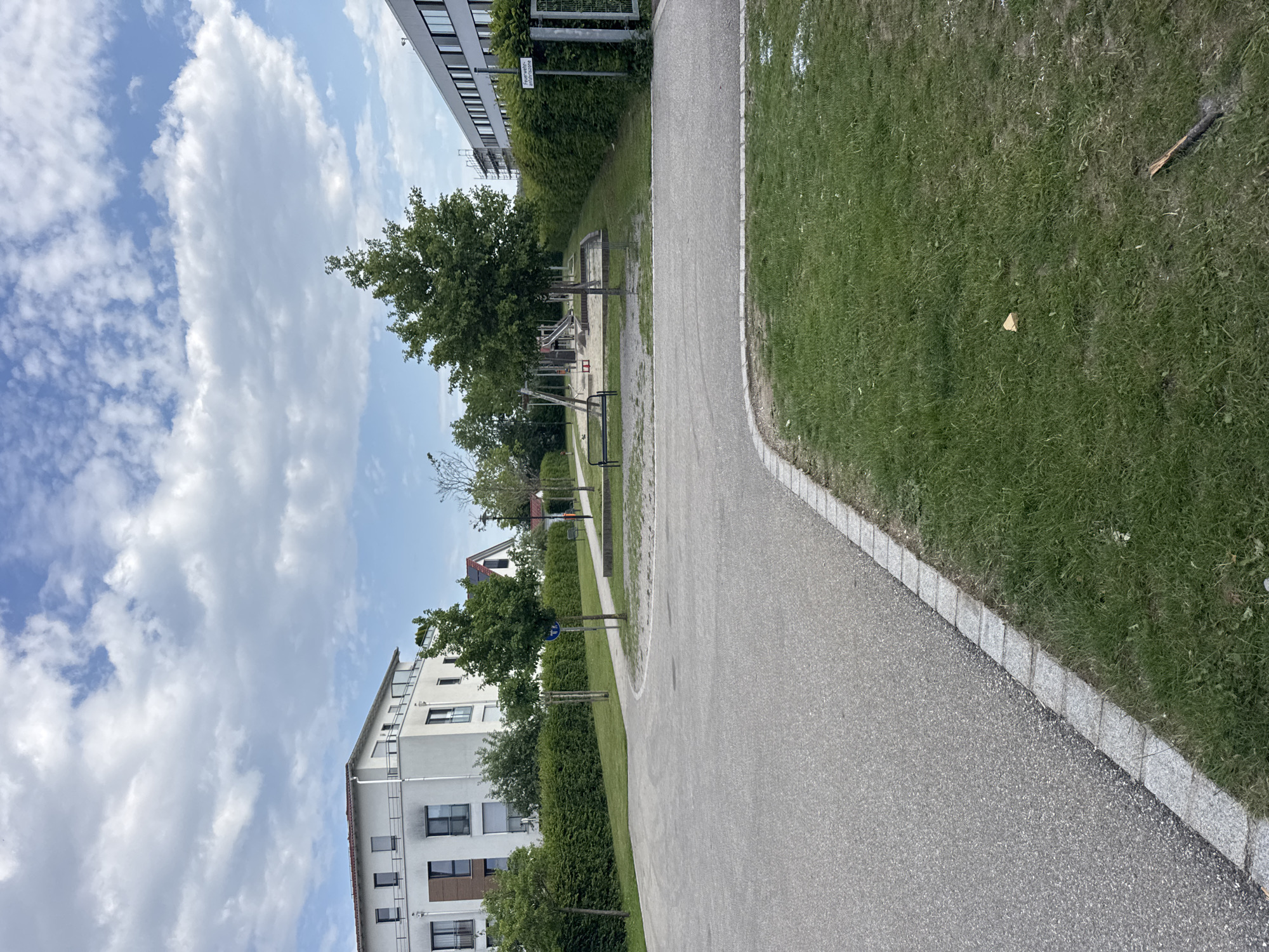 The image shows a street scene in Höhenkirchen-Siegertsbrunn, Germany. In the foreground, a paved road curves to the left, bordered by a strip of green grass and a cobblestone edge. Beyond the road, a well-maintained hedge runs along the right side of the image, separating the road from a children's playground. The playground features a sandpit with a swing set and a small slide. A wooden bench is placed near the sandpit.
Further back, several modern, multi-story buildings are visible. To the left, a white building with multiple windows and balconies is partially obscured by trees. To the right, another building with a gray facade and numerous windows is seen behind the hedge and a fence. The sky above is partly cloudy with scattered white clouds against a blue backdrop. The overall impression is a clean and orderly residential area with recreational facilities.
