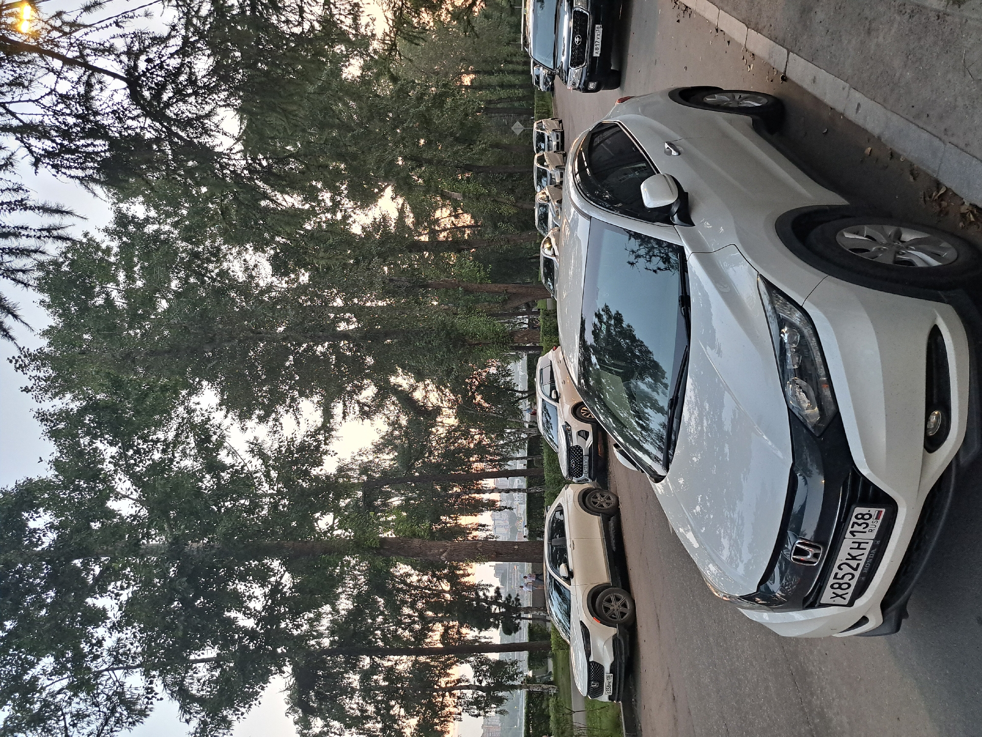 The image depicts a street scene in Irkutsk, Russia, with several cars parked along the side of the road. In the foreground, a white Honda HR-V is prominently featured, its license plate reading "X852KH 138". To its left, another white car, possibly a Honda CR-V, is parked, with its license plate visible as "E358PK 138". Further down the street, a black Toyota Land Cruiser is parked, displaying the license plate "A817YX 38". Several other vehicles are parked in the background, including a white Hyundai Creta. The street is lined with tall trees, and a sidewalk is visible on the right side of the image. In the distance, a body of water and buildings can be seen, suggesting the presence of a city park or waterfront area. The lighting suggests it is either early morning or late afternoon.