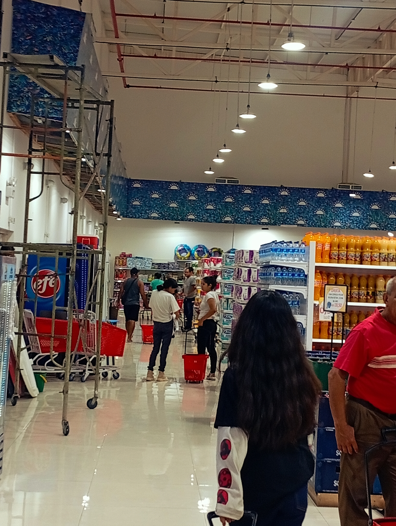 The image shows the interior of a brightly lit supermarket, likely located in Juan Griego, Venezuela, as indicated by the context. The scene captures a wide aisle with a polished, reflective floor. Various shelving units display a range of products, including bottled beverages, packaged goods, and potentially cleaning supplies. In the foreground, a person with long, dark, wavy hair is seen from behind, wearing a black t-shirt over a white long-sleeved shirt adorned with circular, red and black patterns. This individual is holding the handle of a shopping basket.
Further down the aisle, other shoppers are visible. To the left, a person in a white t-shirt and dark pants walks away from the camera. To their right, another individual in a dark tank top and shorts is also walking. In the background, more people are browsing the shelves, and several red shopping carts are present, some empty and some loaded with merchandise. The ceiling features exposed piping and a series of industrial-looking lights. Decorative blue banners with a repeating sun pattern are mounted high on the walls, suggesting a themed decor. The overall impression is that of a typical grocery shopping environment with moderate customer traffic.