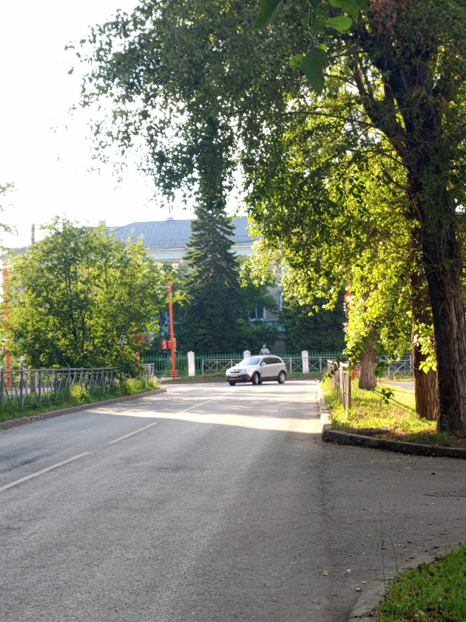 A silver hatchback car is driving down a paved road in Yurga, Russia, on a sunny day. The road has white lane markings and is bordered by a metal fence and grassy areas. Large trees with lush green leaves overhang the road, casting shadows and dappled sunlight. In the background, a light-blue building with a gray roof is visible behind a fence and more trees. Orange poles with traffic signals are also present. The scene suggests a calm, ordinary day in a town with significant greenery. No people are clearly visible, and the car appears to be the only moving object of note. The lighting indicates it is likely late afternoon, given the warm hues and long shadows.