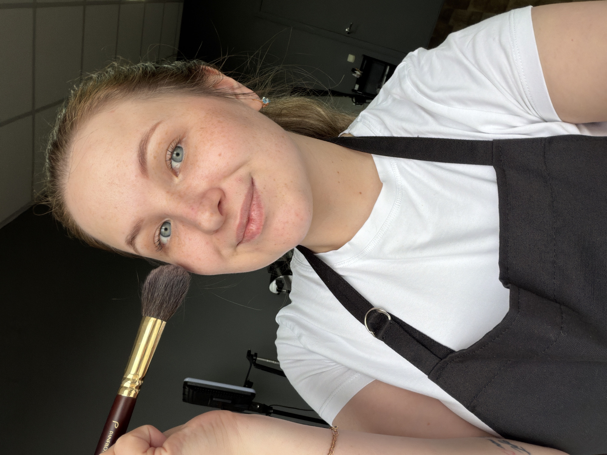 A fair-skinned woman with freckles and blue eyes holds a large, fluffy makeup brush with a brown handle and gold ferrule close to her face. She wears a white t-shirt and a black apron with adjustable straps. Her hair is pulled back. A bracelet is visible on her wrist. In the background is a dark gray wall and metal file cabinets, along with a lighting setup on a stand. The brush is labelled "Panfilo." The location is Chaikovsky, Russia.