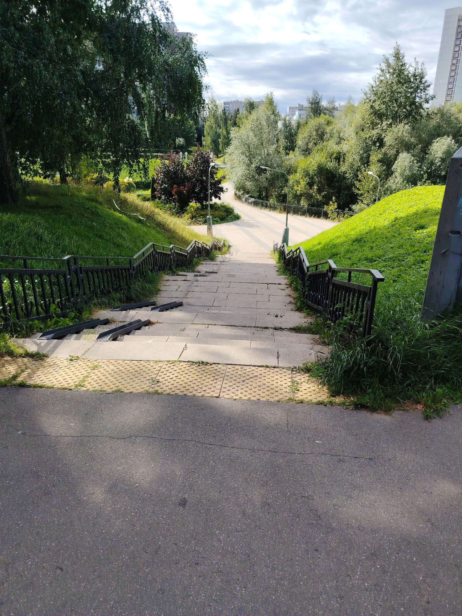 The image shows a descending staircase with metal railings on both sides, leading from a paved area down into a park-like setting. The stairs are made of stone or concrete, with some sections appearing worn. Integrated metal ramps are present on some of the steps, likely for accessibility. The area at the top of the stairs is a paved walkway with yellow tactile paving. To the left and right of the stairs, the ground slopes upwards, covered in green grass and trees. In the background, more trees and buildings are visible, including a tall, modern building on the right side of the image. The sky is partly cloudy. The location is identified as Zelenograd, Russia, and the scene appears to be a public outdoor space, possibly a pedestrian underpass or pathway within a residential or park area.