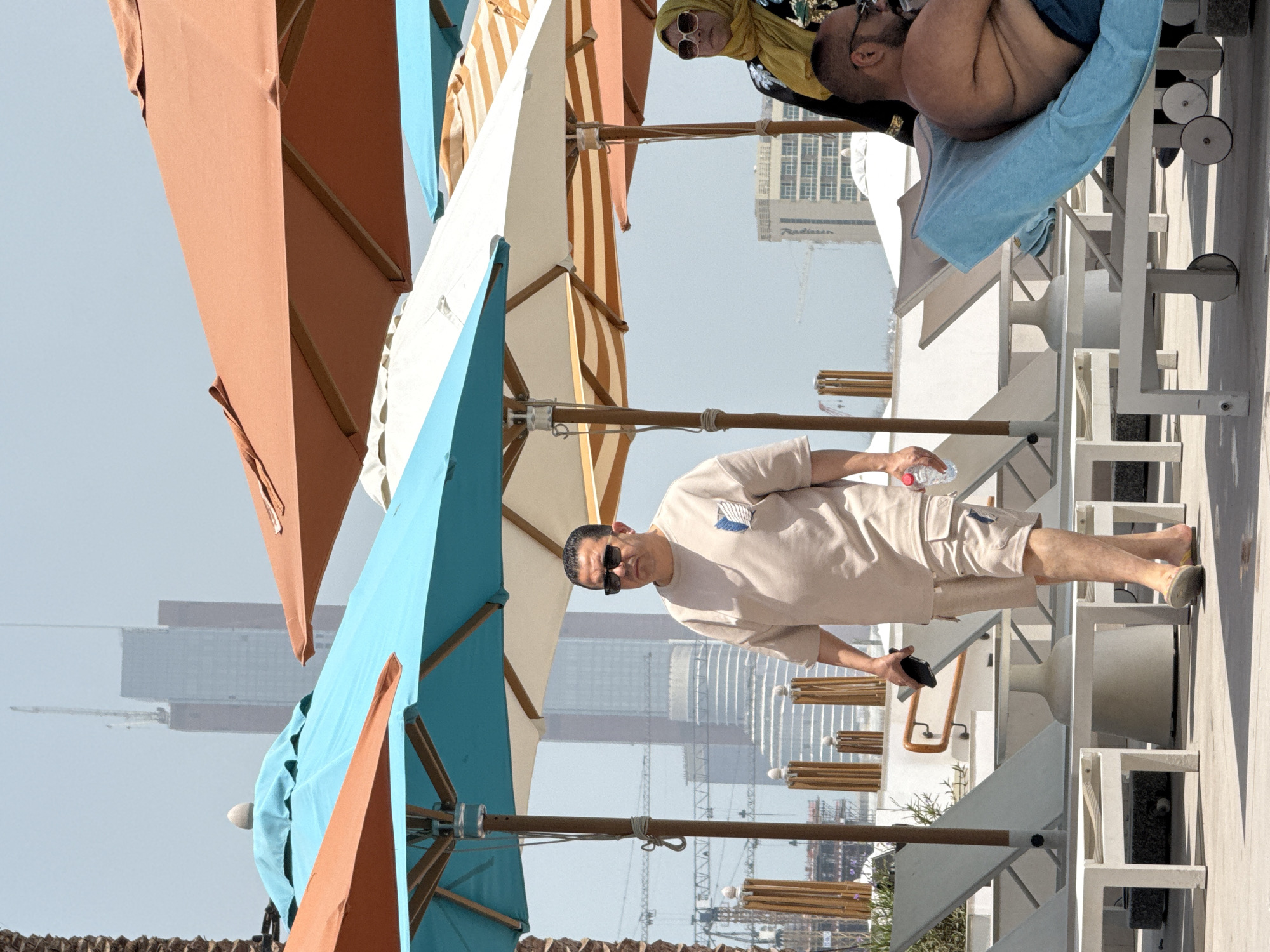 The image depicts an outdoor scene with several people and umbrellas under a bright sky, likely in a resort or public area. In the foreground, a man in a light-colored t-shirt and shorts walks across a paved area. He is wearing sunglasses and carrying a mobile phone in his left hand and a bottle of water in his right. Behind him, a woman in a yellow hijab and sunglasses is visible, partially obscured by umbrellas. To the right, another man is reclining on a lounge chair, his torso bare.
Several lounge chairs are arranged on the pavement, some with white frames and others with a light blue covering. A collection of large umbrellas, in colors such as turquoise, orange, and striped beige and orange, are positioned throughout the scene, providing shade. In the background, skyscrapers and construction cranes suggest an urban environment. The overall impression is a sunny day at a recreational facility, possibly a pool or beach club, within a city.