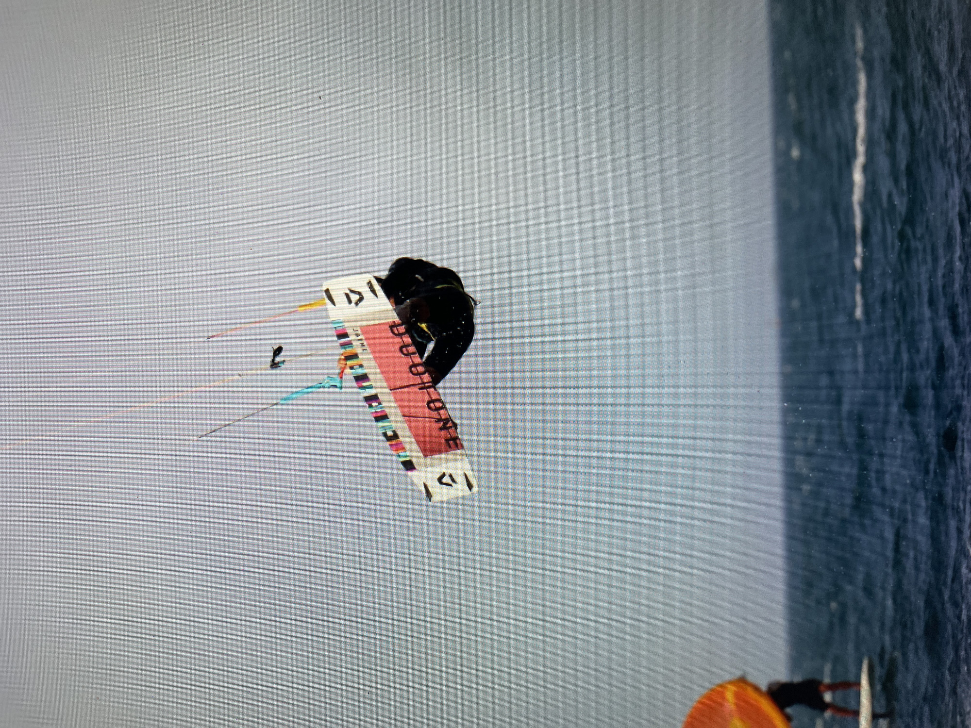 The image shows a person kitesurfing in Bacong, Philippines. The person is wearing a black top and is attached to a kiteboard labeled "DUOTONE" with colorful stripes. The kiteboard is in the air, with the kite lines visible extending towards the left side of the image. The background shows a clear sky above and the ocean on the right side. Another person wearing an orange life vest is standing in the water near the lower right corner of the image.