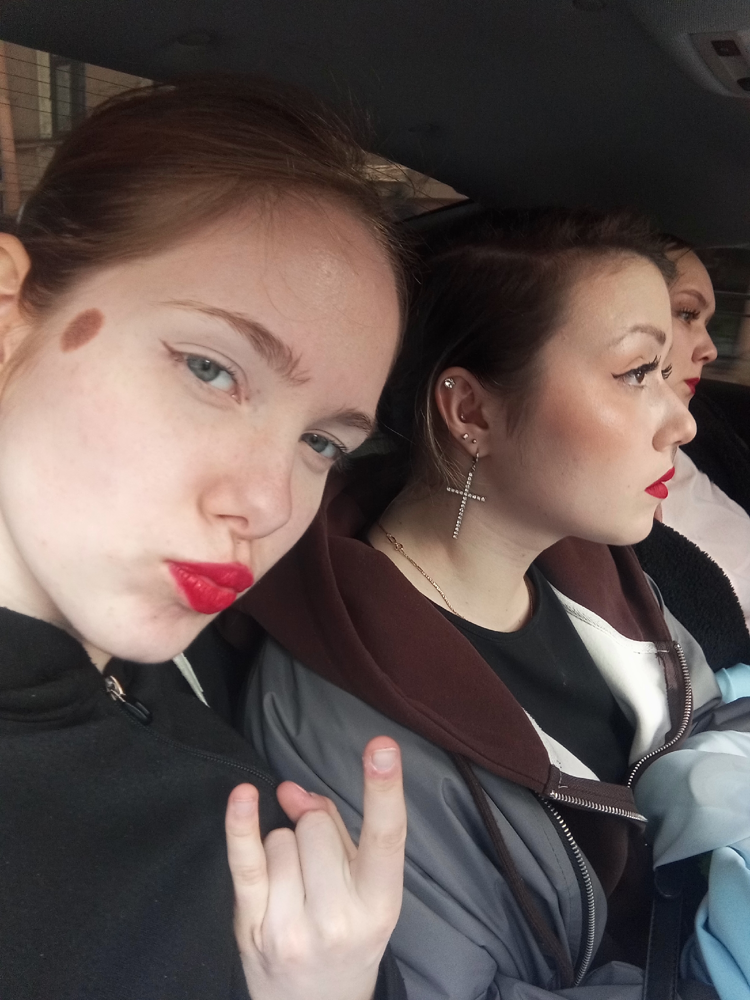 Three individuals are in a vehicle, likely a car, based on the overhead. The lighting suggests an indoor or shaded outdoor environment.
The person in the foreground, on the left, is a young woman with fair skin, reddish-brown hair, and a mole on her forehead. She is wearing a black zip-up hoodie and has bright red lipstick. She is making a "rock on" hand gesture.
The second person, in the middle, is a young woman with dark hair pulled back. She is wearing a large, jeweled cross earring and a gold necklace. She has red lipstick and a dark-colored shirt layered under a gray and maroon jacket. She is looking toward the right.
The third individual, partially visible in the background on the right, is another person with red lipstick and a light-colored shirt.
The environment appears to be the interior of a vehicle. Based on the architecture faintly visible through the windows, and that the user requested Saint Petersburg, it is possible that this image was taken somewhere in the city of Saint Petersburg, Russia. However, there are no other cues or identifying features that concretely indicate this location.