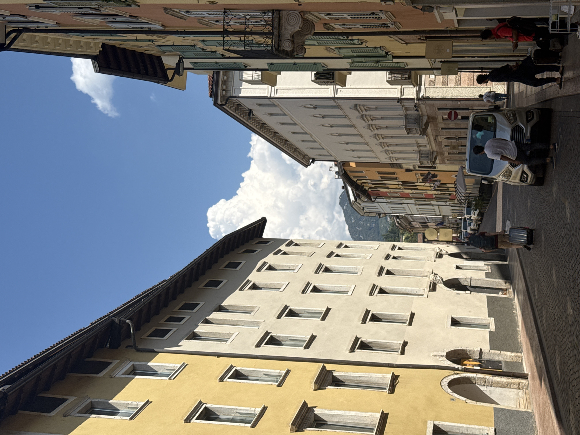 The image depicts a sun-drenched cobblestone street in the city of Trento, Italy, lined with tall, colorful buildings. The architecture is characterized by traditional European styles, with multiple stories, numerous windows, and decorative facades. On the left, a building with a bright yellow facade and arched entrances dominates the foreground. Adjacent to it is a lighter-colored building with a more muted tone. On the right, buildings display a range of colors, including orange and yellow, with green shutters and ornate balconies.
The sky is a vibrant blue, dotted with fluffy white clouds, suggesting a clear and pleasant day. In the background, mountains are visible, hinting at the natural landscape surrounding Trento.
Several people are present on the street, engaged in various activities. In the mid-ground, a person is pulling a rolling suitcase, suggesting travel or arrival. Further down the street, a silver van is parked, and several individuals are standing near it or walking along the sidewalk. One person is wearing a backpack.
The street itself is paved with cobblestones, and shadows are cast by the buildings, indicating the strong sunlight. The overall impression is a lively urban scene in a picturesque Italian city.