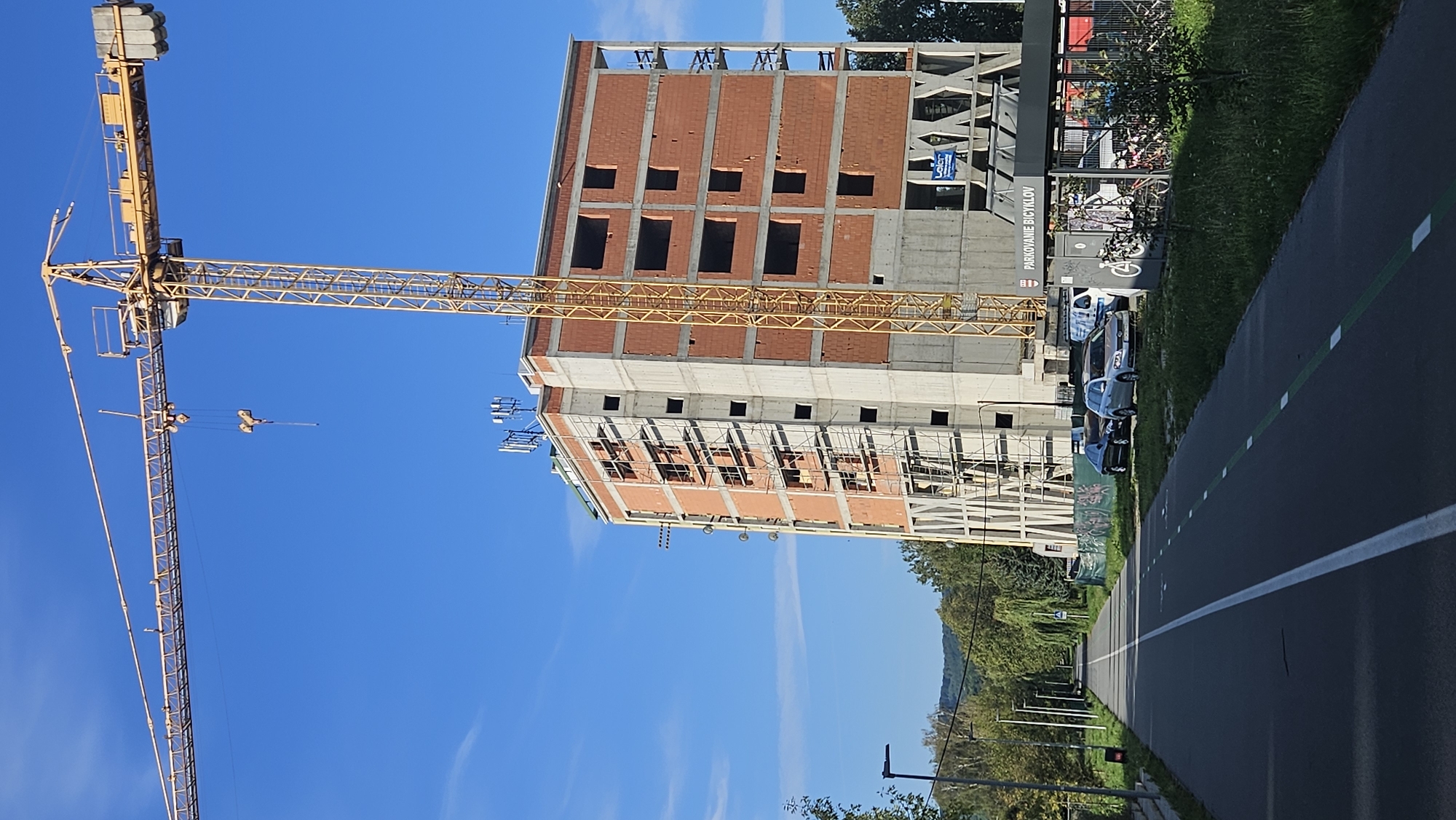 The image depicts a construction site in Prešov, Slovakia, featuring a tall, unfinished building and a yellow tower crane. The building, a multi-story structure with exposed brickwork and concrete elements, is partially clad in scaffolding. The tower crane is positioned alongside the building, with its jib extending horizontally across the frame. In the foreground, a paved pathway, marked with white and green lines, runs alongside a grassy area. A bike rack with bicycles is visible near the pathway, adjacent to the construction site. Several cars are parked near the bike rack. The background shows trees and hills under a clear blue sky. The overall scene suggests ongoing construction work in an urban or semi-urban environment.