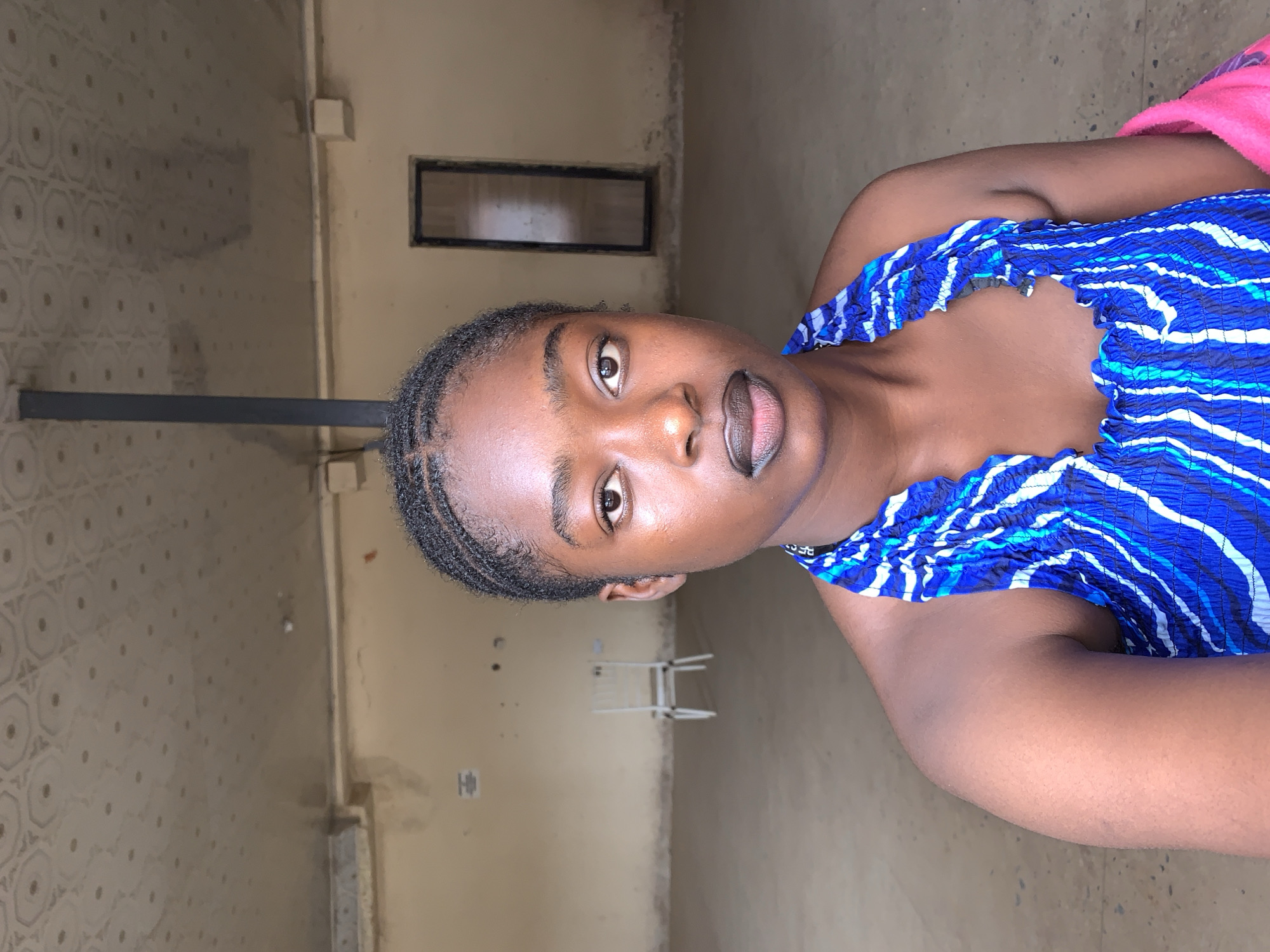 A young, dark-skinned woman is indoors, facing the camera. She wears a blue and white patterned strapless top and her hair appears to be styled close to her head. The background reveals an interior with pale yellow walls and floor. A dark metal pole extends horizontally from the upper left wall. A rectangular window is visible further back. A white, folded laundry drying rack stands on the floor, further back. The location is consistent with an interior in the New Stadium area of Nigeria, based on the general appearance and objects present.