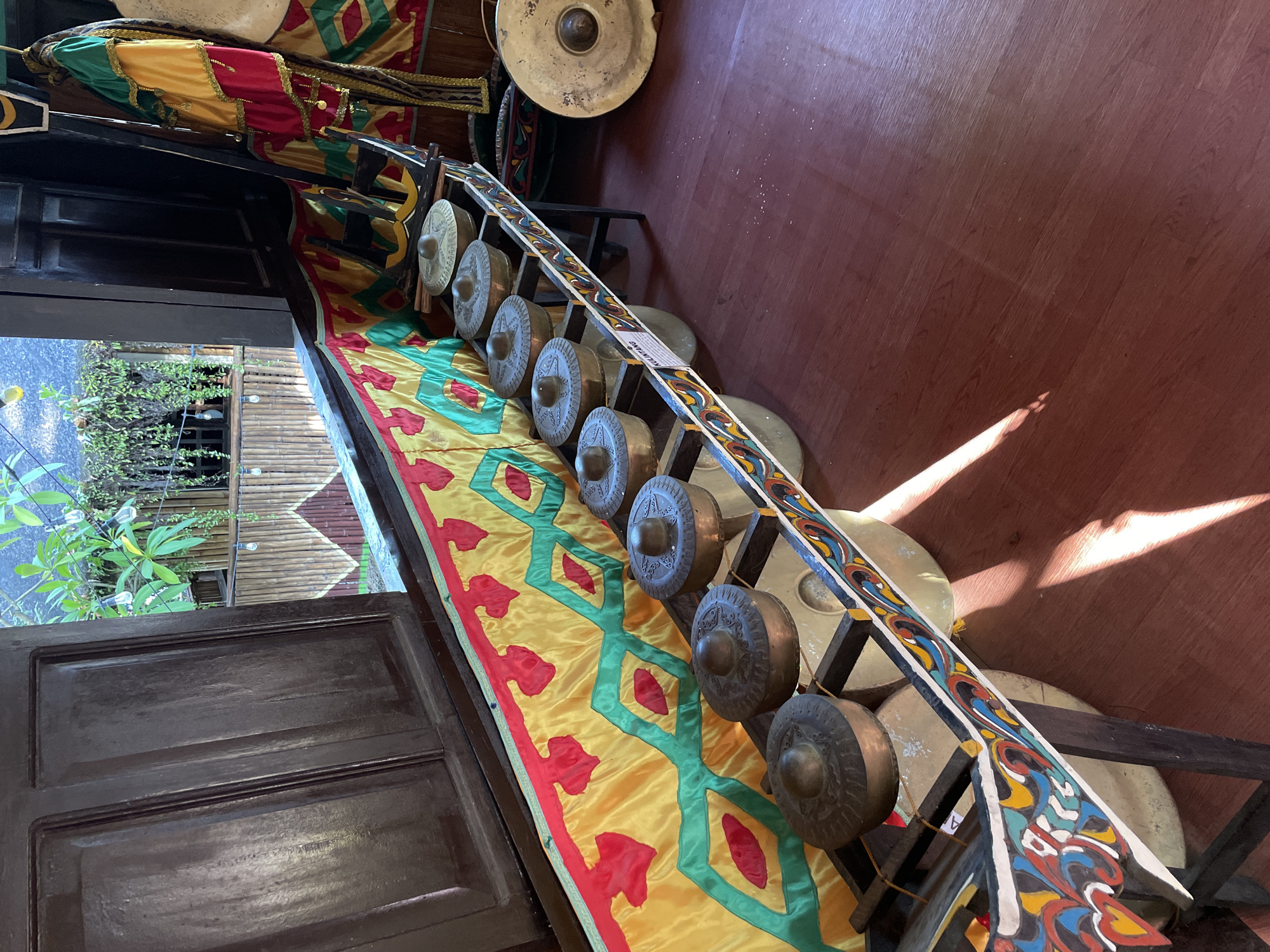 An interior view of a room showcases traditional musical instruments, specifically a kulintang, displayed on a wooden stand. The kulintang, a set of bronze gongs arranged horizontally, is adorned with colorful, intricate patterns. Behind the kulintang, a vibrant fabric with yellow, red, and green patterns drapes along the wall. To the right, another large, flat bronze gong is visible. The floor is made of polished dark wood, reflecting sunlight that streams in from an unseen source. In the background, through an open window, lush green foliage and bamboo structures are visible, suggesting an outdoor or semi-outdoor area. The presence of traditional instruments and cultural decor indicates the location is likely a cultural center, museum, or a traditional house in Davao City, Philippines. No people are present in the image.