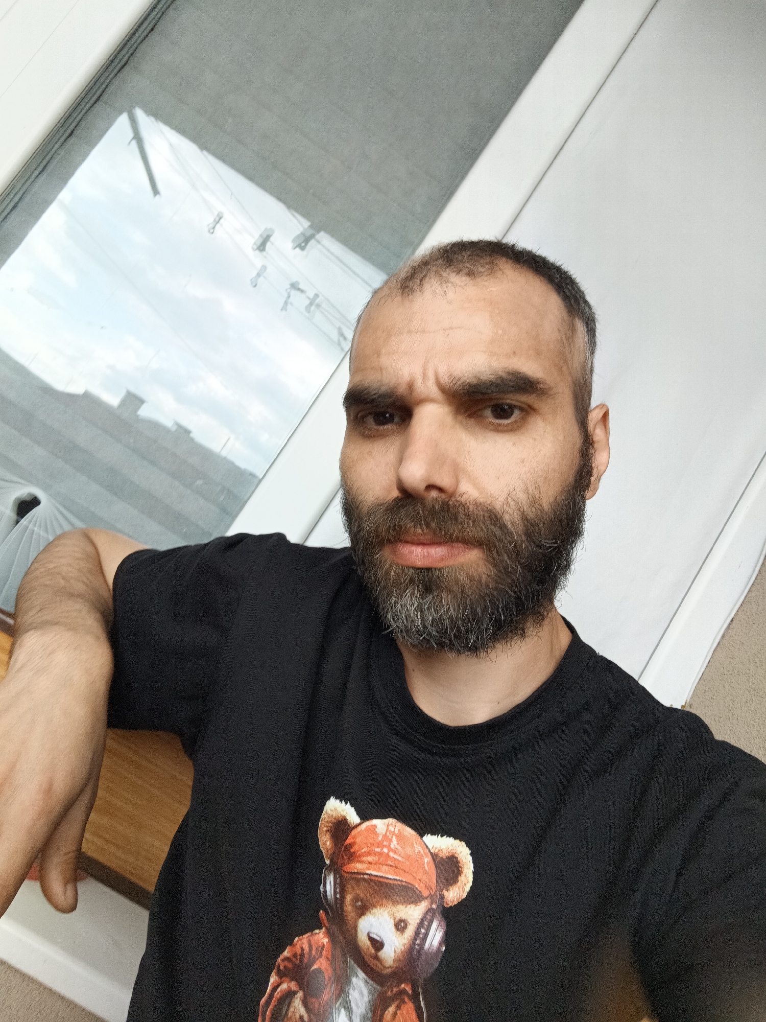 A man with a dark beard and a black t-shirt with a bear print is sitting indoors near a window. The location is Unknown, Unknown.