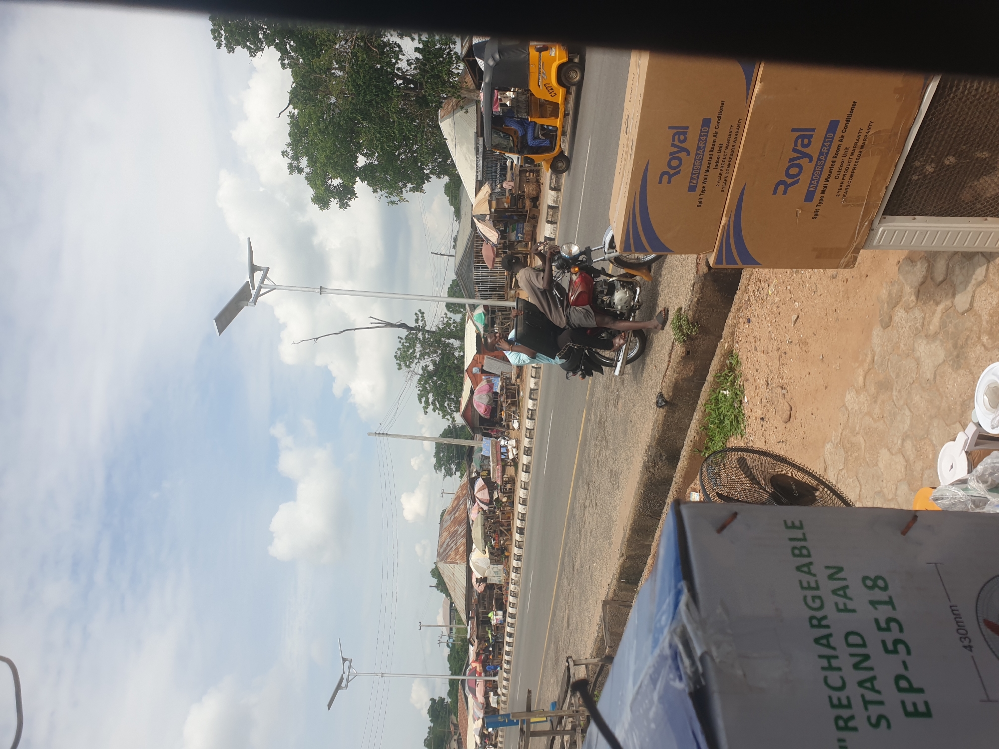 The image, taken in Adankolo, Nigeria, captures a roadside scene. A man sits astride a motorcycle parked along the shoulder of the road. A yellow "Keke" tricycle drives on the road. Open-air market stalls line the roadside. In the foreground are boxes labeled "Royal," indicating air conditioners, and a box for a "RECHARGEABLE STAND FAN." Solar-powered streetlights are visible along the road. The sky above is partly cloudy.