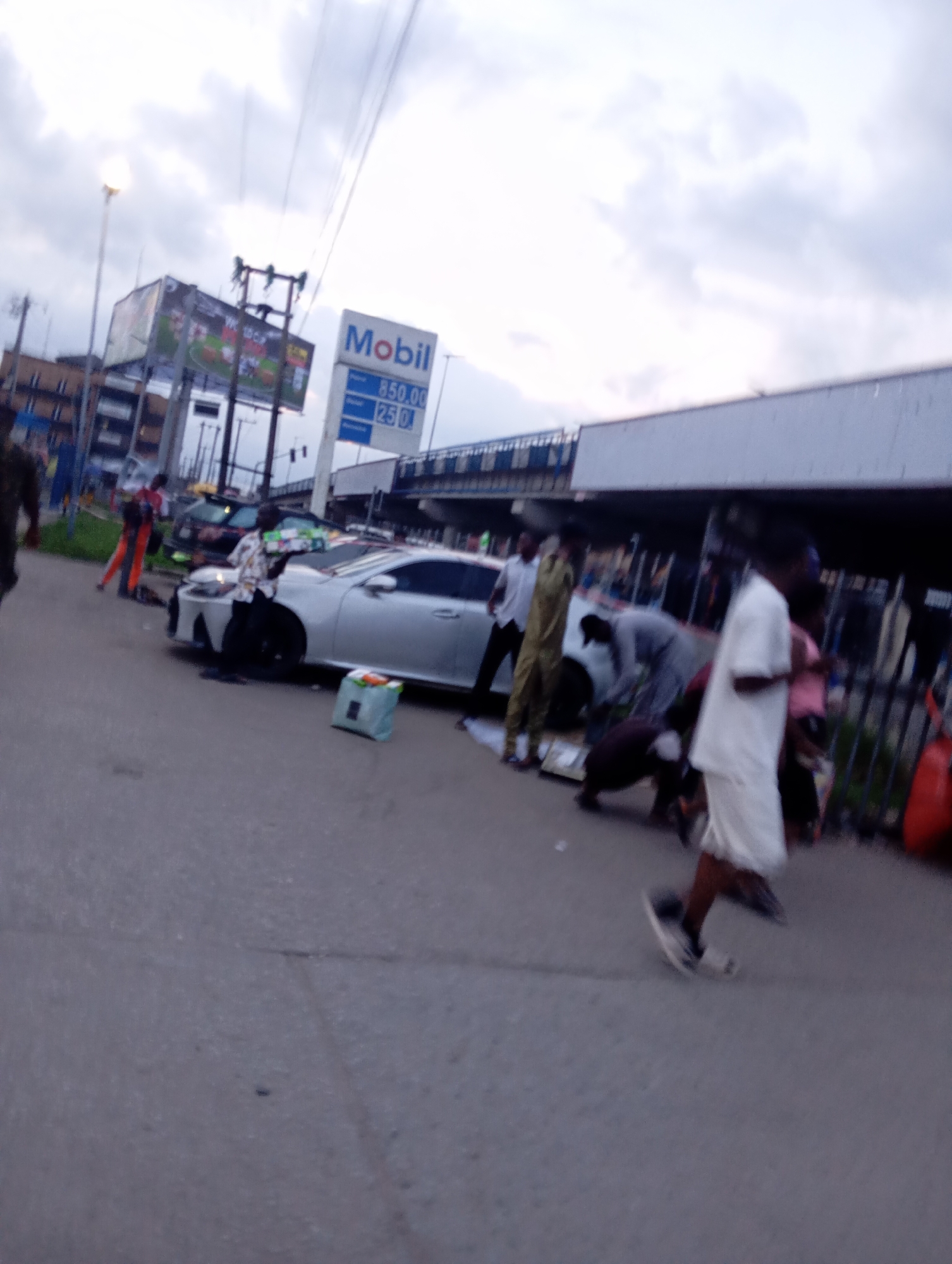 The image depicts an outdoor scene on a street in Port Harcourt, Nigeria. A silver sedan is parked on the right side of the road. Several individuals are gathered around the car, some appear to be engaged in activities related to the vehicle or its contents. A person in the foreground, wearing a white shirt and shorts, walks away from the camera. In the background, a Mobil gas station sign is visible, indicating the presence of a fueling station. The sky is overcast, suggesting a cloudy day. The overall impression is of a busy street scene with commercial activity.