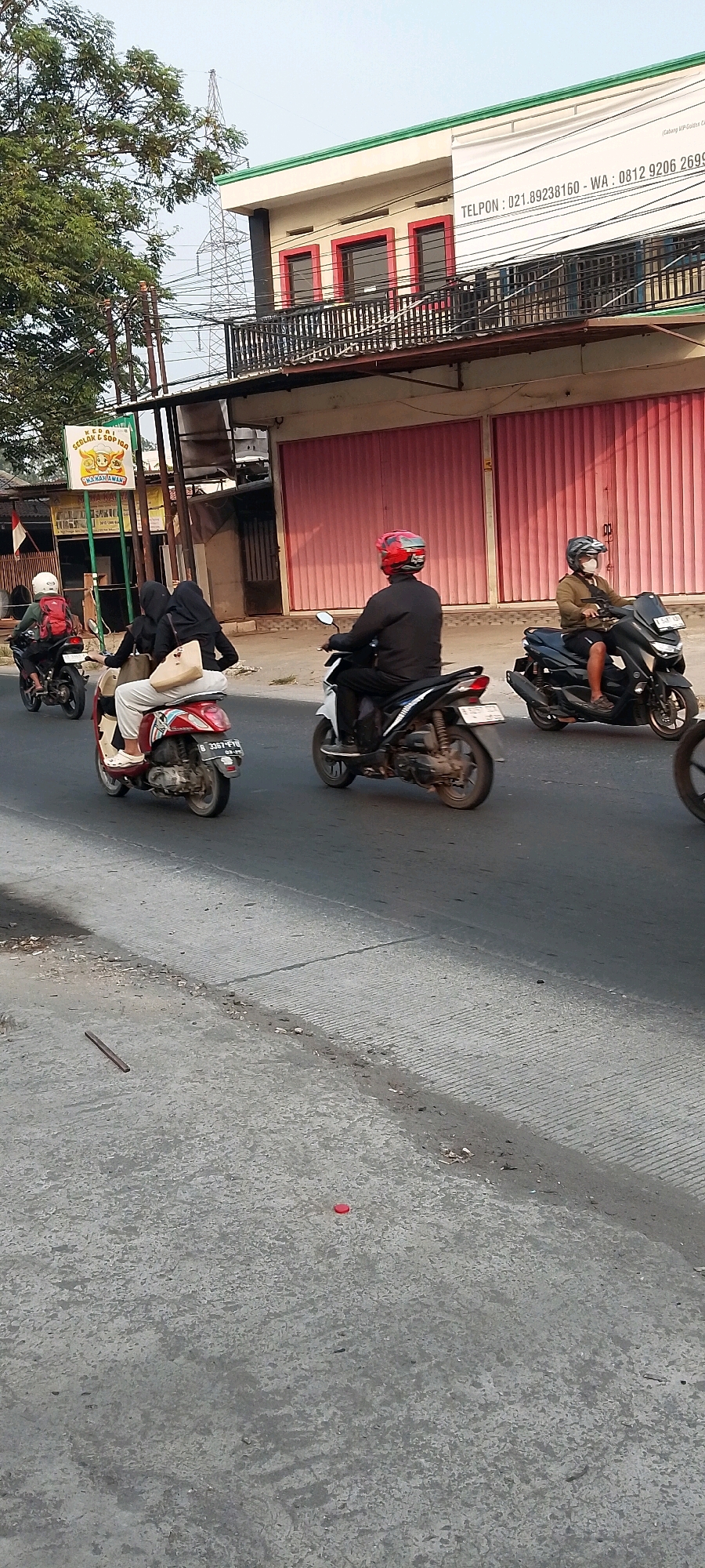The image depicts a street scene in Satriamekar, Indonesia, on a clear day. Several people are riding motorcycles. In the foreground, a person wearing a black jacket and a red helmet is riding a white motorcycle. To their right, another person wearing a light brown jacket and a face mask is riding a dark blue motorcycle. Further in the background, a person in a white helmet and red backpack is riding a motorcycle on the left side of the road. On the left side of the road, two people, one wearing a white shirt and the other in a black hijab, are riding a red scooter. They are carrying a light brown bag.
The environment includes a road with gray asphalt, cracked in some areas. On the left side of the road, there is a building with a green trim and red window frames, featuring a banner with white text. A large tree with green foliage is also visible on the left. The background shows more buildings and utility poles. A small red object, possibly a bottle cap, is on the road in the lower middle of the image.