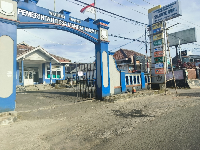 The image shows the entrance to the "Pemerintah Desa Mandalamukti" (Village Government of Mandalamukti) in Cikalong, Indonesia. It is a blue, arched gate with the sign "Pemerintah Desa Mandalamukti" inscribed on it. The gate is adorned with a small Indonesian flag on the right side. There is a single-story building behind the gate with windows and a door, likely serving as the village administration office. On the right side of the gate, a wooden ladder is leaning against a signpost featuring various signs with Indonesian text. There is an asphalt road in front of the gate. The sky is clear and blue with wispy white clouds.