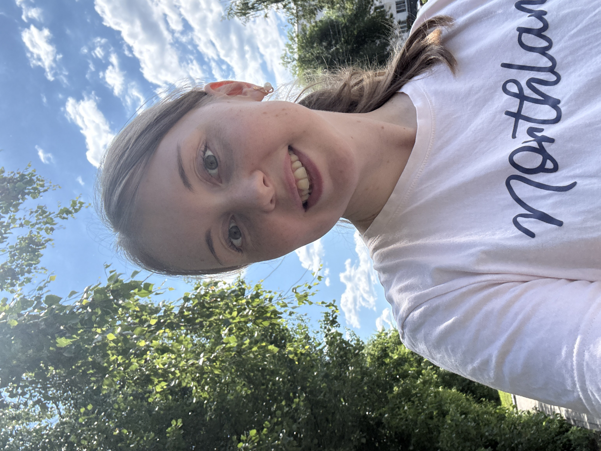 An outdoor, waist-up, eye-level, medium-shot selfie shows a Caucasian woman with light skin and freckles, smiling directly at the camera. She has light brown hair pulled back into a ponytail, and she is wearing a light pink t-shirt with the word "Northland" written in navy blue cursive. The background is a bright blue sky filled with fluffy white clouds, with the green leaves of a tree on the left side of the frame and the tops of some buildings and trees on the right. The sunlight is bright, casting shadows and creating a lens flare in the upper left quadrant.