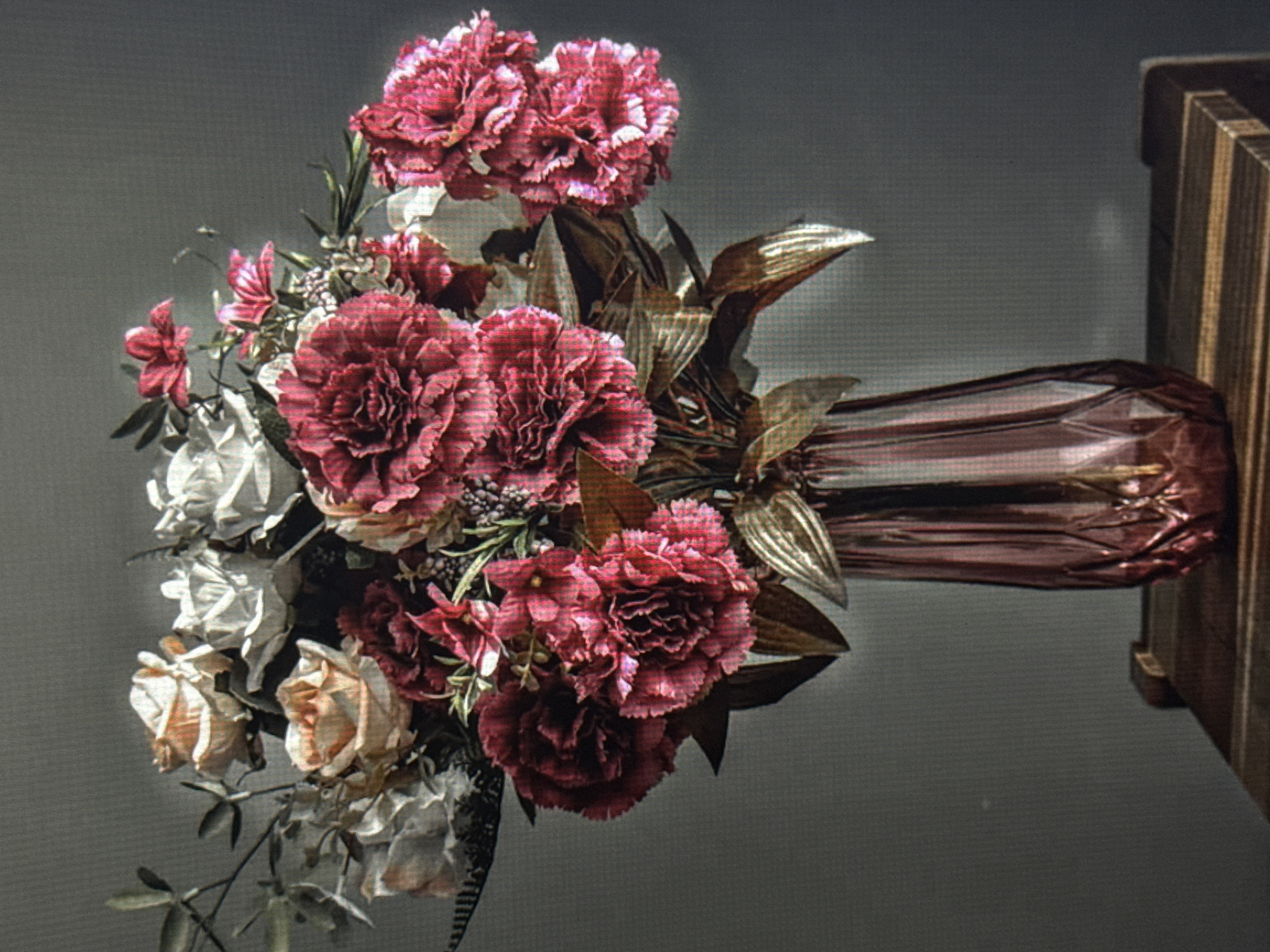 This media file features a close-up, rotated shot of a floral arrangement and a vase. The arrangement consists of several large, ruffled pink and crimson carnations, alongside lighter pink and white roses, with some greenery and small buds. The flowers are densely packed, creating a lush bouquet.
The vase is elongated and appears to be made of a glossy, metallic material with a reddish-brown hue. Its surface has a crinkled or textured appearance, reflecting light in places. The vase is positioned to the right of the flowers, and the bouquet is mostly to its left. Behind the arrangement and vase, the background is a plain, dark gray, which helps the flowers and vase stand out. To the far right of the image, a portion of a wooden object, possibly a stand or piece of furniture, is visible. The lighting is somewhat dim, casting soft shadows and highlighting the textures of the flowers and vase. There is no discernible text in the image, nor any indicators of time of day, weather, or specific emotions. The overall scene suggests a still-life composition, likely indoors, focused on the beauty of the flowers and the decorative vase.