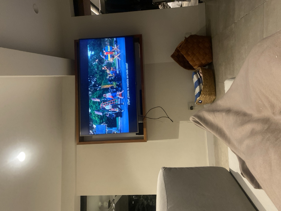 A television mounted on the wall displays a scene with people, flags, and equipment, with the dialogue "Oye! ¿Cómo te metiste en todo esto?" visible at the side. Two baskets, one woven and one holding a striped item, sit on the wall. A section of the room includes a couch partially covered by a light brown blanket.