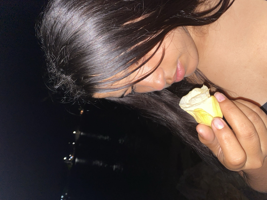 The image captures a close-up, angled profile shot of a person, likely a woman, with long, dark, straight hair. The subject is positioned in the upper right portion of the frame, with her head tilted downwards and slightly to the left. Her dark hair cascades around her face and shoulder, reflecting some light, indicating its sheen.
Her face is partially visible in profile, showing her forehead, eyebrow, a prominent nose, and full, naturally colored lips that appear slightly parted. Her skin tone is medium to dark. She is holding a single, pale yellow or off-white flower petal or a small, unopened flower bud in her right hand, bringing it delicately close to her nose, suggesting she is smelling or gently observing it. Her eyes are not clearly visible, but her downward gaze implies concentration or contemplation. Her nails are neatly manicured and appear to have a natural or light pink polish.
The background is very dark, almost black, signifying nighttime. In the distant lower-left portion of the image, faint, blurred lights are visible, some appearing as streaks or reflections, which could suggest a body of water reflecting distant city lights or lights from other objects in the environment. The overall atmosphere conveyed is one of quietude, introspection, or a moment of appreciation for the flower. No text is visible in the image.