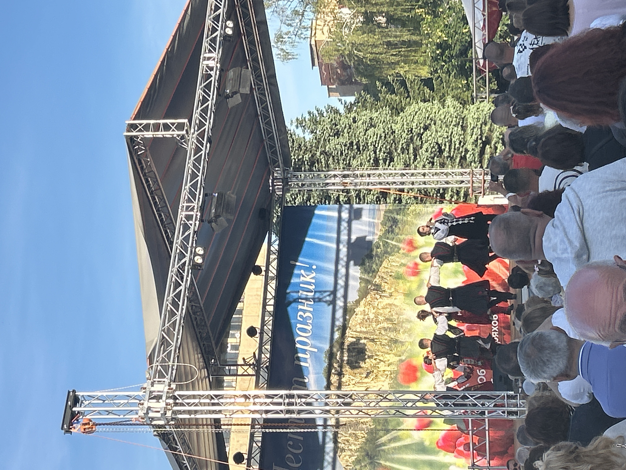 The image depicts an outdoor performance in Gorna Oryahovitsa, Bulgaria. A stage with metal scaffolding supports a backdrop showing a landscape with red poppies. A group of dancers in traditional black and white costumes are performing. A crowd of people of mixed ages and ethnicities watch the performance from ground level. Buildings and trees are visible in the background under a blue sky.