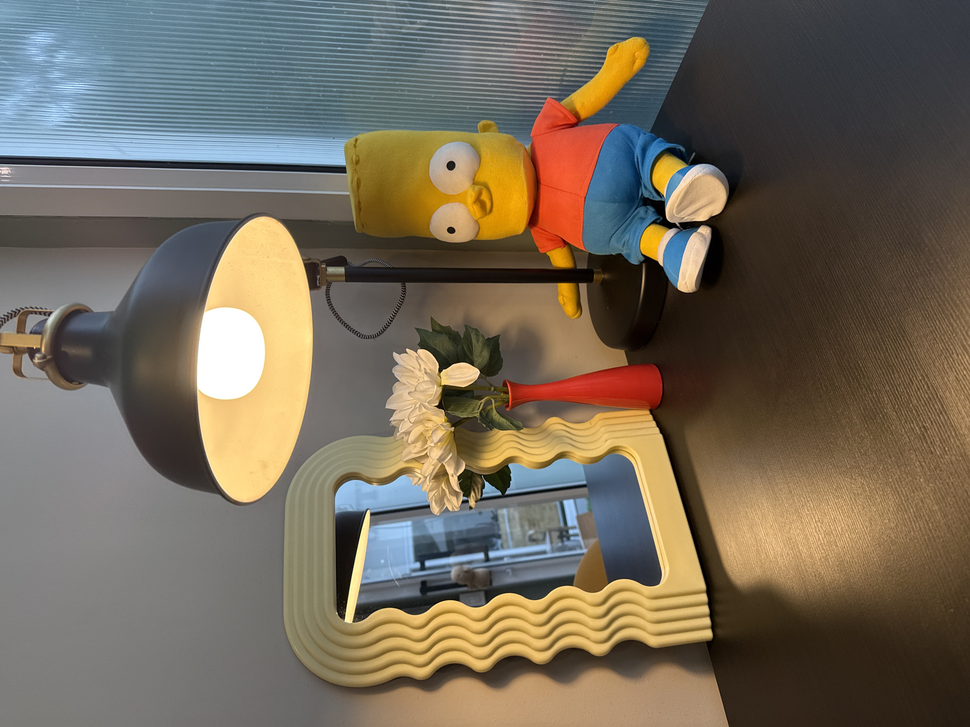 The image shows a Bart Simpson plush toy sitting on a dark wooden desk next to a red vase containing white artificial flowers. A yellow, wavy-edged mirror is behind the flowers. A black desk lamp with a bright bulb is positioned to the left of the plush toy, casting a warm glow on the scene. The background features a light gray wall and a fluted glass window. There are no people visible in the image. The scene is likely indoors, possibly in a home or office setting, with no specific indicators pointing to Bangkok, Thailand.