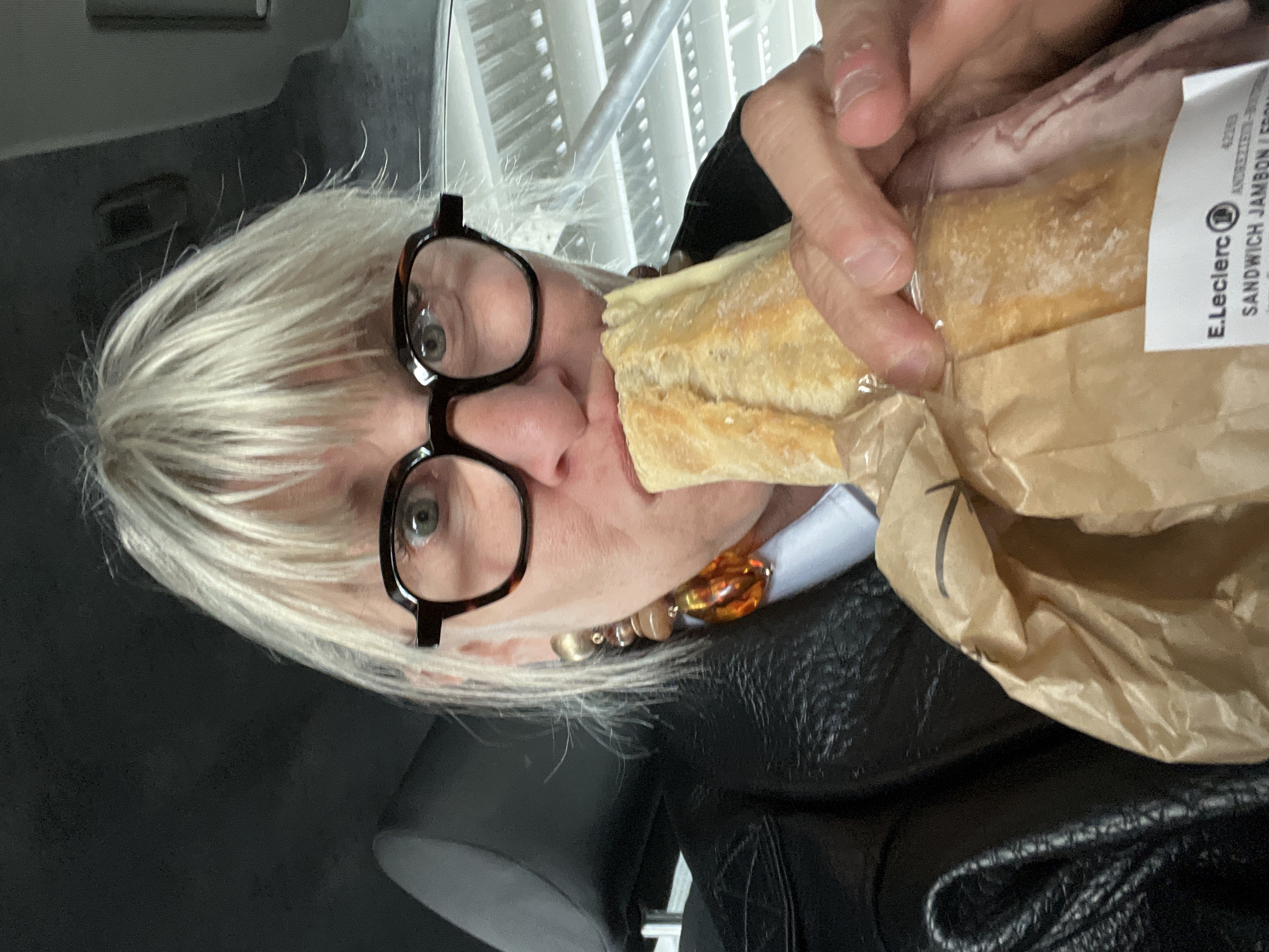 A person with short, blonde hair and eyeglasses is eating a sandwich. The sandwich appears to be a "Sandwich Jambon Lerdon," according to the label from "E.Leclerc." The location of the sandwich appears to be in Andrézieux-Bouthéon, France. The person is wearing a black jacket, a white shirt, and a necklace with amber-colored beads. They are holding the sandwich in a paper bag. The background includes a window with horizontal blinds. The person may be in a car.