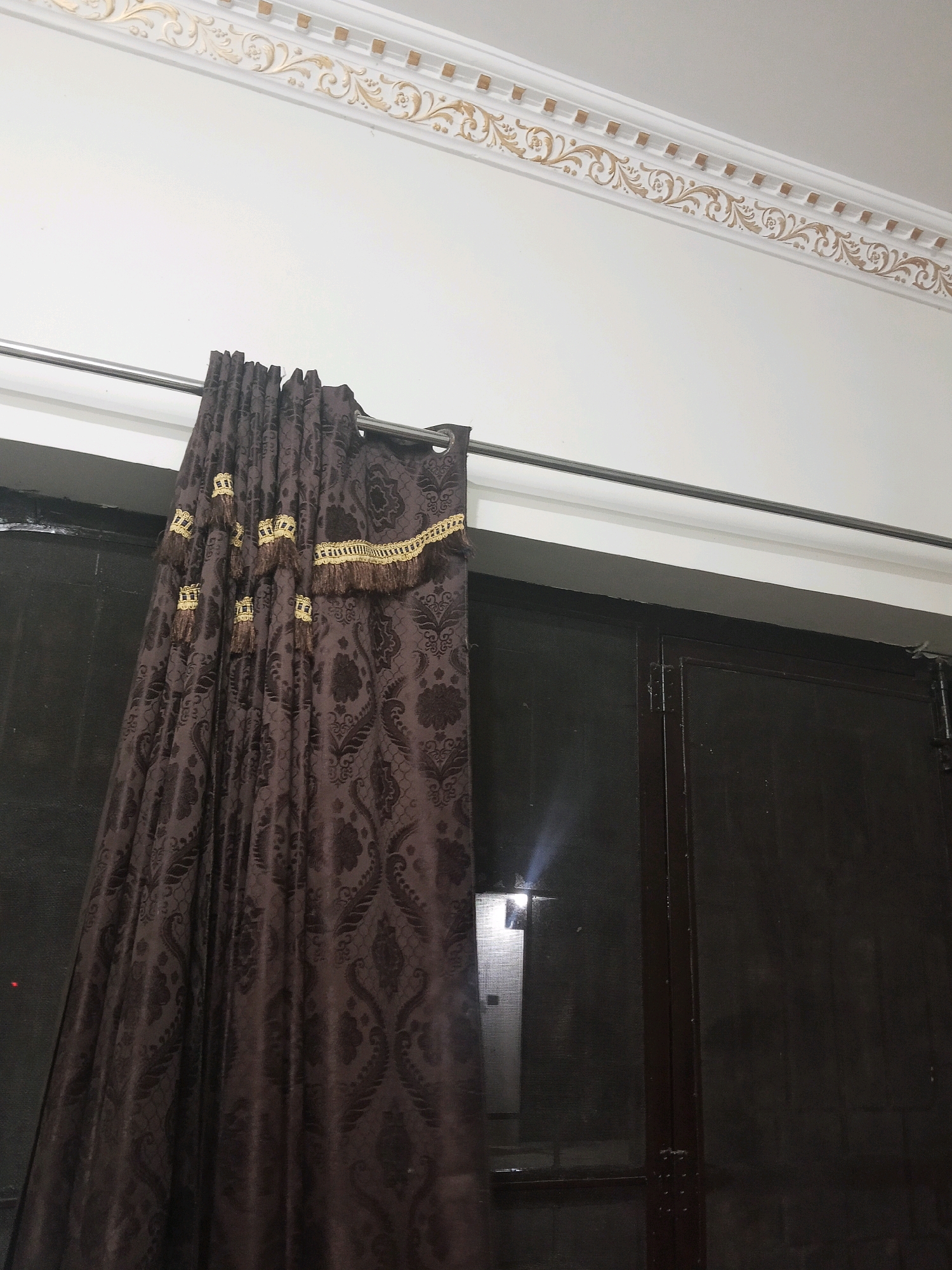 The image shows a close-up view of a brown curtain with a damask pattern, hanging from a silver curtain rod. The curtain is embellished with gold trim and tassels that resemble crowns. Above the curtain, a white ceiling with ornate gold molding is visible. The curtain appears to be covering a dark window or door. The scene is indoors, with lighting suggesting it may be nighttime or a dimly lit room. The location is identified as Bhedian - Nai Basti, Pakistan.