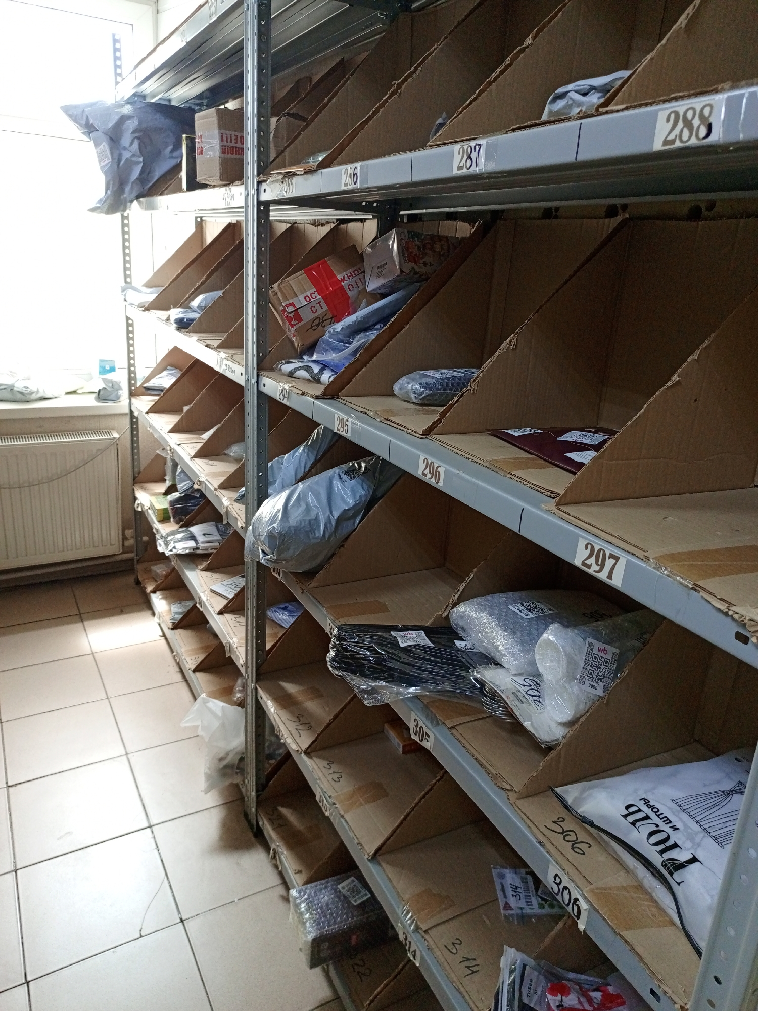 The image shows a shelving unit, likely in a stockroom or storage area. The shelves are metal and divided into sections, each holding various packages and items. The sections are labeled with numbers, ranging from 286 to 314, indicating a systematic organization. The packages vary in size and shape, some wrapped in plastic or bubble wrap, while others are in cardboard boxes. The lighting is somewhat dim, and the background includes a tiled floor and a radiator near a window. Overall, the scene depicts an organized storage space with packaged items ready for distribution.