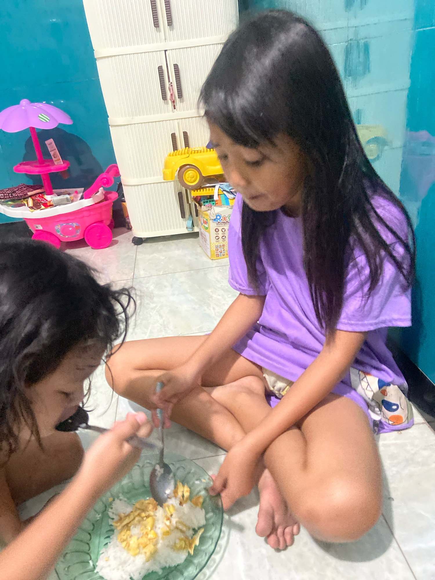 Two young girls are seated on a tiled floor in what appears to be a home. The girl on the right, wearing a purple t-shirt, is facing forward and looking down at a plate of food. She has long black hair. The girl on the left, partially obscured, is actively eating from a green plate filled with rice and scrambled eggs. She is also using a spoon to eat. In the background, there is a tall white cabinet with multiple drawers, a pink toy shopping cart with an umbrella, and other toys. A yellow toy vehicle is visible on top of the cabinet. The walls are a bright teal color. The scene suggests a casual mealtime in a domestic setting, possibly in Gelam, Indonesia, given the context.