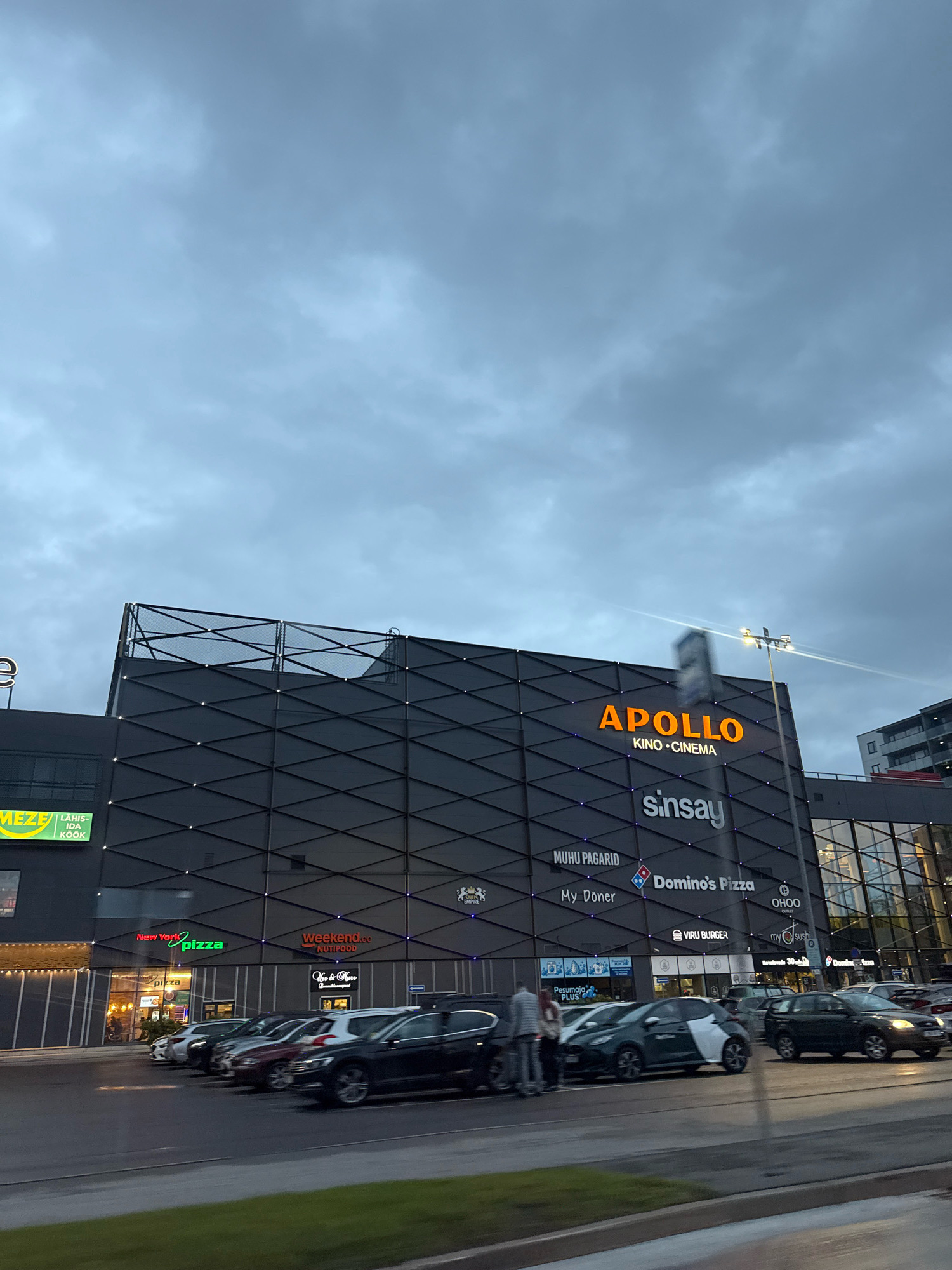 The image shows the Apollo Kino building in Tallinn, Estonia, on a cloudy day. The building is modern, with a dark facade decorated with a geometric pattern of diamond shapes. The "APOLLO KINO CINEMA" sign is prominently displayed in orange neon letters. Below it, several other businesses are visible, including "sinsay," "MUHU PAGARID," "My Doner," "Domino's Pizza," "OHOO GRILL," "New York Pizza," and "weekend.ee NUTIPOOD." The lower portion of the building features large windows and storefronts. In front of the building, a street with several parked cars and a few people walking is visible. The sky is overcast, with dark gray clouds. The overall impression is of a modern urban commercial and entertainment center.