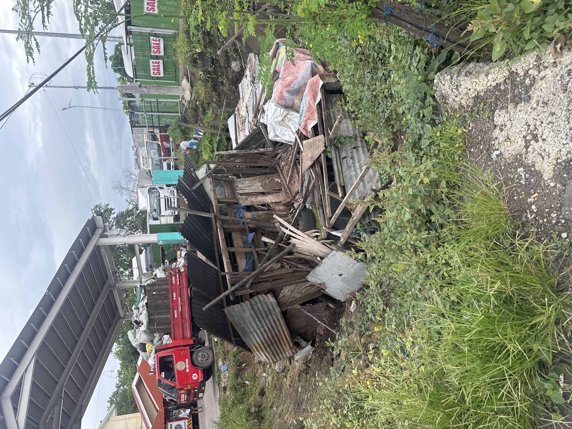 The image depicts a roadside scene in Davao City, Philippines. A pile of debris, consisting of corrugated metal, wood scraps, and tarpaulin, sits among overgrown vegetation on the right side of the frame. A red dump truck, filled with white sacks of material, is visible on the left side, positioned under a covered structure. In the background, trucks are parked near a green building with "SALE" signs. The sky is overcast. People are present in the background near the red truck.