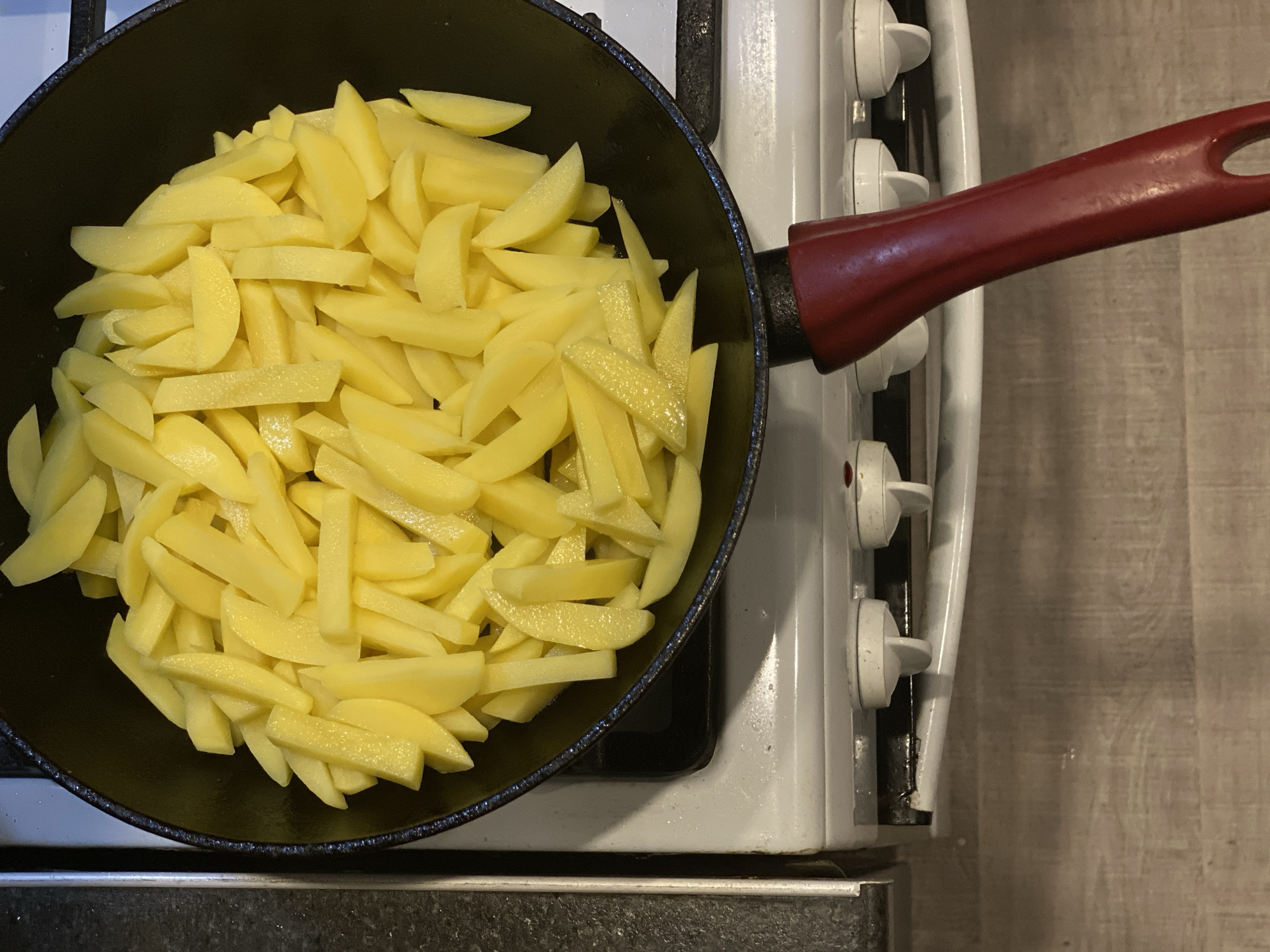 A top-down, medium shot depicts a black frying pan filled with raw, cut potatoes on a white gas stove. The pan has a red handle. The stovetop is slightly dirty, showing some wear and tear. The background includes a countertop with what appears to be a black and gray pattern and a light wood-like surface. The location appears to be a domestic kitchen, likely within a residential dwelling. No people or specific actions are evident. The scene portrays a preparation stage of cooking. There is no indication or evidence that places this scene in Sochi, Russia.