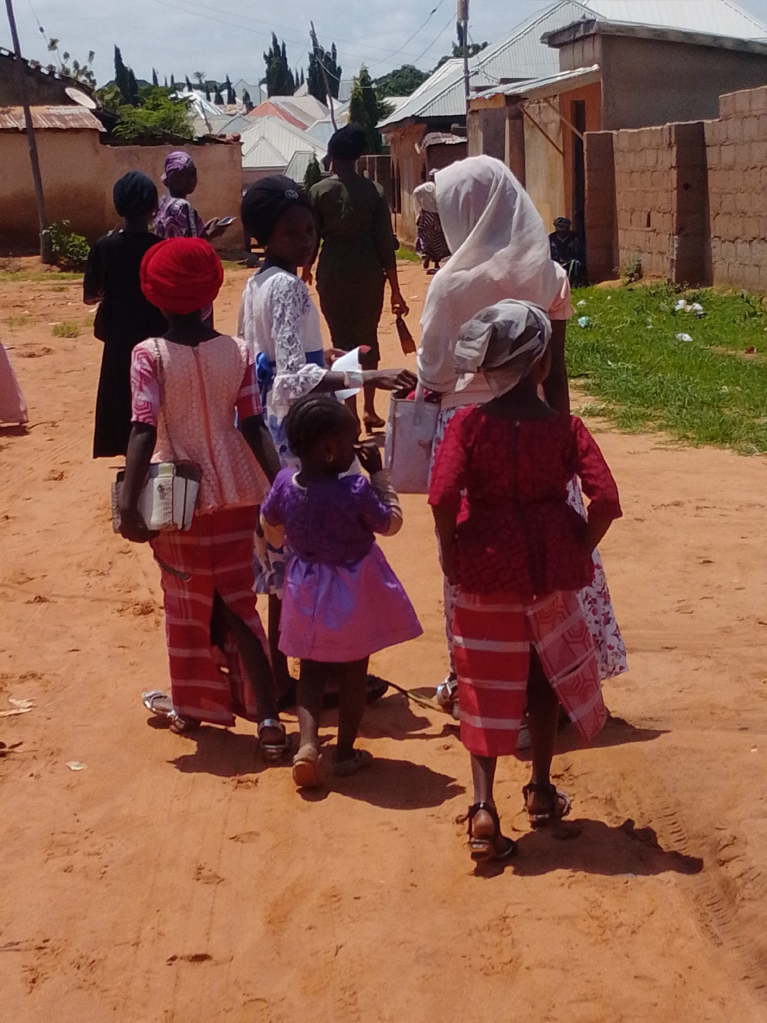 The image depicts a group of people, primarily women and girls, standing and walking on a dirt road in Gombe, Nigeria. The scene appears to be in a residential area with buildings made of brick and corrugated iron roofs visible in the background. The sky is overcast.
In the foreground, several individuals are captured from behind, showcasing their attire. One person is wearing a bright red headscarf and a patterned light pink peplum top with a long, striped red and white skirt. They are holding a white object that resembles a book or folder. Next to them, a young girl in a purple dress is also facing away from the camera. Further to the right, another individual is wearing a red patterned top and a similar striped skirt, their hands clasped behind their back.
Other individuals are visible in the middle ground and background. One woman wears a white hijab and a light-colored garment. Another person is dressed in a dark green outfit, and a woman in the background is wearing a purple and white striped dress. Some people are walking away from the camera, while others appear to be stationary. The ground is reddish-brown dirt, with patches of green grass and some scattered debris on the right side of the frame. Footprints and tire tracks are visible in the dirt.