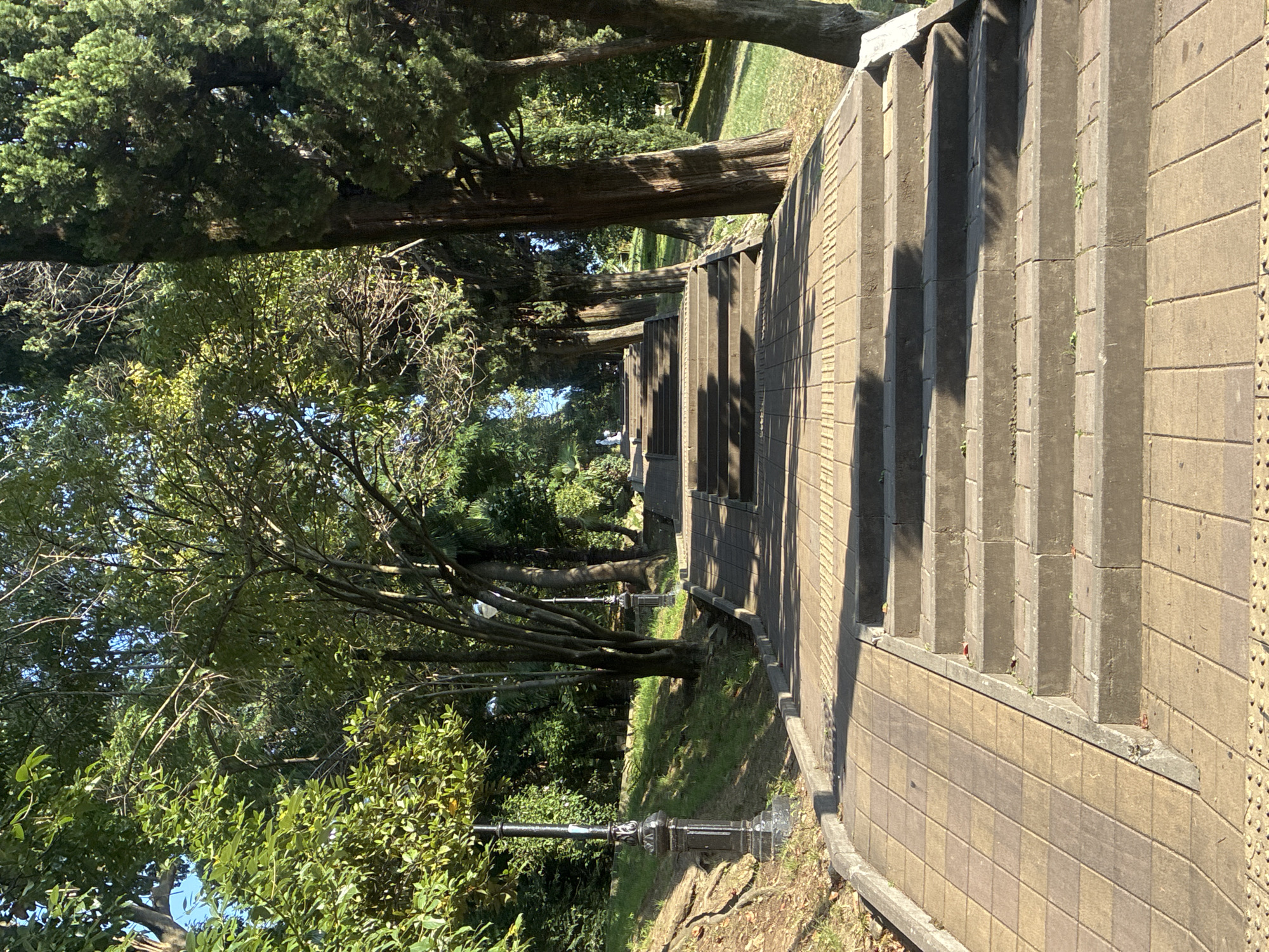 The image shows a stone staircase ascending a hill in a park-like setting. The staircase is bordered by a paved walkway and a ramp on the left side. Mature trees with dense green foliage line both sides of the staircase, creating a shaded canopy. Sunlight filters through the leaves, casting dappled shadows on the steps and walkway. In the distance, at the top of the staircase, a white statue is visible. A lamppost with an ornate base stands on the left side of the walkway near the bottom of the stairs. The scene appears to be in an outdoor public space, likely a park or garden, with the natural elements of trees and vegetation dominating the view. The presence of the statue suggests it may be a memorial or decorative feature. The overall impression is one of a tranquil, well-maintained public area. The location is identified as a park in Sochi, Russia, a city known for its subtropical climate and numerous parks and recreational areas.