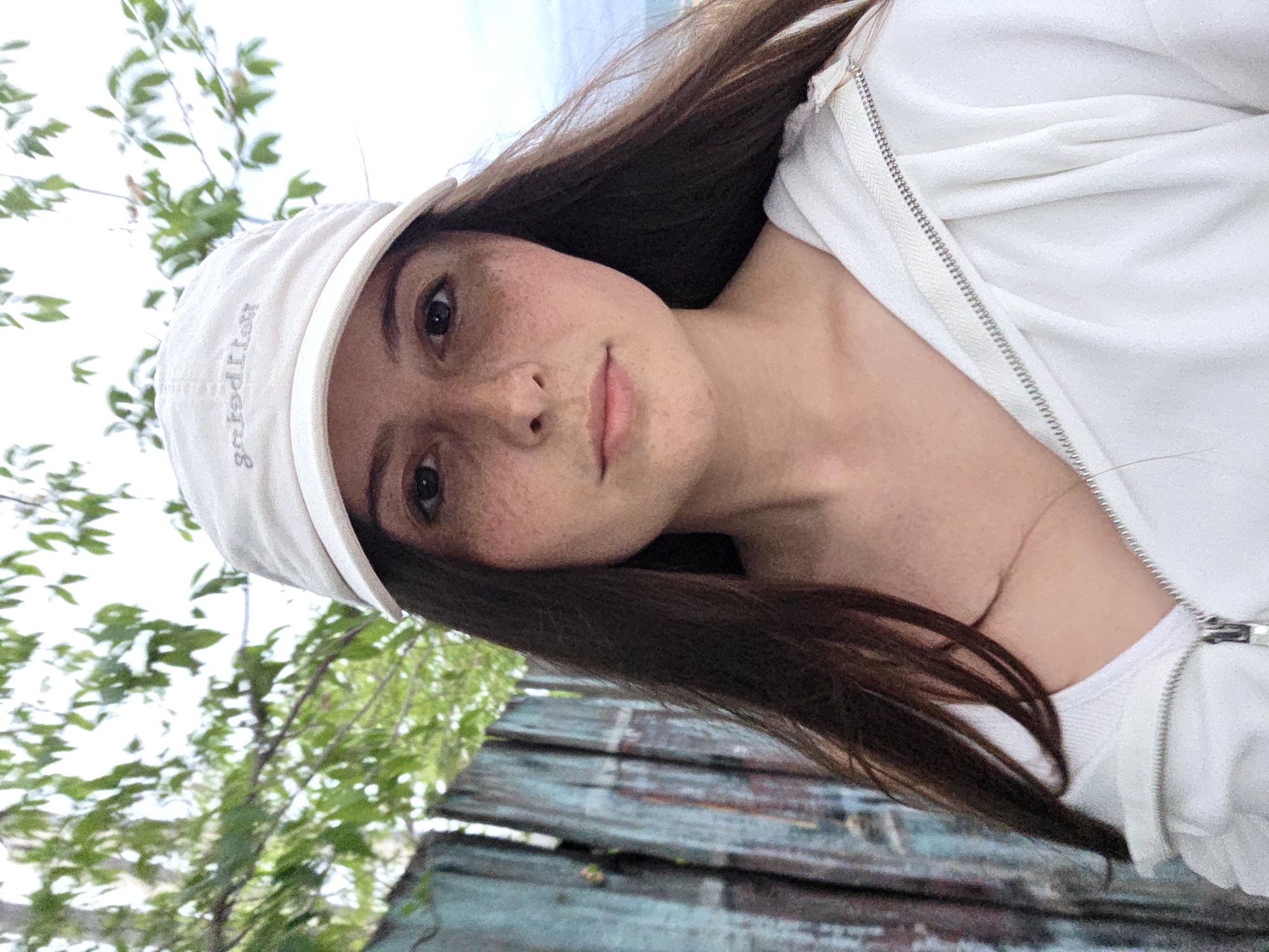 A young woman with long, dark hair and freckles is pictured from the chest up, facing forward. She is wearing a white baseball cap with embroidered text, a white zip-up hoodie, and a white V-neck shirt underneath. The background features a weathered, turquoise-painted wooden fence and green foliage. The sky above is overcast. The image appears to have been taken outdoors.