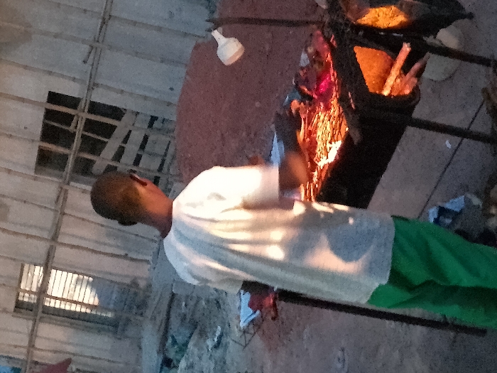 The image depicts a scene at what appears to be dusk or night, centered around a person and a cooking fire. The photograph is oriented vertically, rotated 90 degrees clockwise from a standard upright perspective.
In the immediate foreground, a person stands facing slightly away from the camera. They have short, dark hair and are wearing a light-colored (likely white or off-white) short-sleeved shirt or t-shirt and green trousers. The warm, orange light from a nearby fire illuminates their back and right side, creating highlights on their clothing.
To the right of the person, a robust cooking setup is visible. A large, dark-colored pot or pan rests on a sturdy metal stand. Beneath the pot, a vivid fire blazes, casting bright orange and red light, serving as the primary illumination for this part of the scene. Inside the pot, a yellowish substance, possibly food like rice or grain, can be seen. Adjacent to the fire, what appears to be fuel, such as wooden sticks or logs, is stacked, ready to be added to the flames.
The ground surrounding the cooking area seems to be rough and unpaved, likely dirt or an uneven, natural surface. In the background, extending above and to the left of the person, a rudimentary overhead structure is apparent. It consists of a grid-like arrangement of multiple wooden beams or planks, suggesting a temporary shelter, scaffolding, or an unfinished building. A rectangular dark opening or window is visible within this structure. A distinct white, bulb-shaped object is seen hanging in the upper right corner of the frame, which could be a lamp or part of the structure.
The overall lighting is dominated by the warm glow of the fire, which creates strong contrasts and deep shadows in the areas it doesn't directly illuminate. The scene conveys an impression of outdoor or semi-outdoor cooking activity taking place during the evening or nighttime hours. No discernible text is visible within the image.