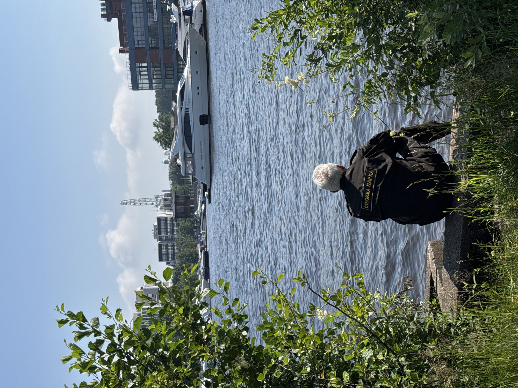 A person wearing a black uniform with a yellow patch that reads "СЛУЖБА ПАРКА" (Park Service) is sitting on a concrete barrier at the edge of a body of water. The person has gray hair and is facing away from the camera, looking out at the water. In the background, across the water, there are several modern buildings, some with large windows, and a Ferris wheel structure. Several yachts are docked in the water. Green foliage frames the foreground and the right side of the image. The sky is blue with scattered white clouds. The scene appears to be a waterfront area in Saint Petersburg, Russia, likely a park or recreational zone.