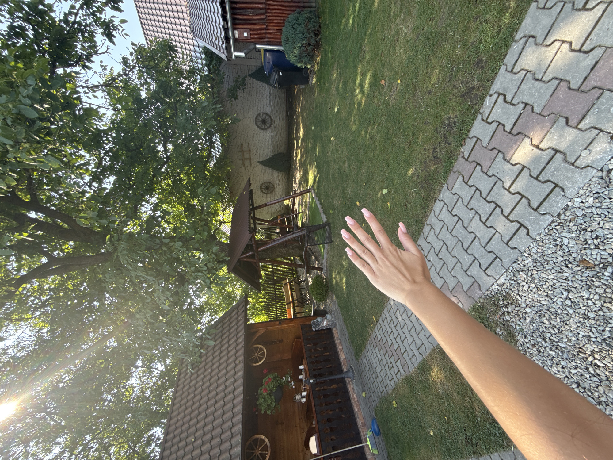 The image captures a sunlit backyard in Cârțișoara, Romania. In the foreground, a person's arm and hand, with painted nails, are visible, extending into the frame from the left. The hand is open, with fingers spread. The background features a grassy lawn, a paved pathway made of interlocking concrete stones, and a gravel area. To the left, a wooden covered patio area with a dark wooden railing and a bench is partially visible, adorned with decorative wagon wheels and hanging flower baskets. Beyond this structure, a wooden swing set with two seats stands under a large tree, its branches creating dappled shadows on the lawn. In the distance, a white brick wall is adorned with two more decorative wagon wheels and two small conical evergreen trees. To the right of the wall, two large trash bins, one black and one blue, are placed near a wooden shed with a corrugated roof. The scene is framed by lush green trees and a clear blue sky, with sunlight streaming through the foliage.