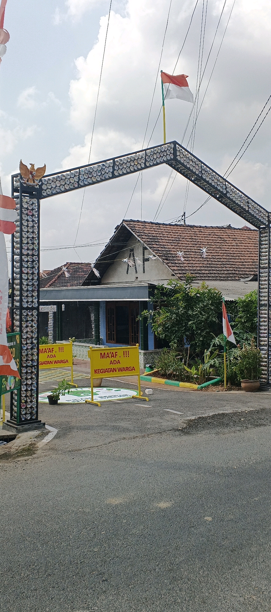 This is a medium shot, outdoor, eye-level, vertical, full-color photograph taken on a cloudy day. The scene depicts an entrance archway made of what appears to be recycled plastic bottles and metal, decorated with reflective material. An Indonesian flag flies atop the arch. To the left of the arch is a vertical banner with Indonesian text and flags. In the background, a single-story house with a tiled roof is visible. Two yellow signs with red lettering stand in the foreground, indicating "MA'AF, !!! ADA KEGIATAN WARGA" (Sorry, there is a community event). A small potted plant is placed in front of the left sign, and the ground inside the archway is painted with a circular design. The road in front of the archway is asphalt, and power lines crisscross the sky above. The overall impression is a temporary entrance or checkpoint for a local event in a residential area of Bluri, Indonesia.