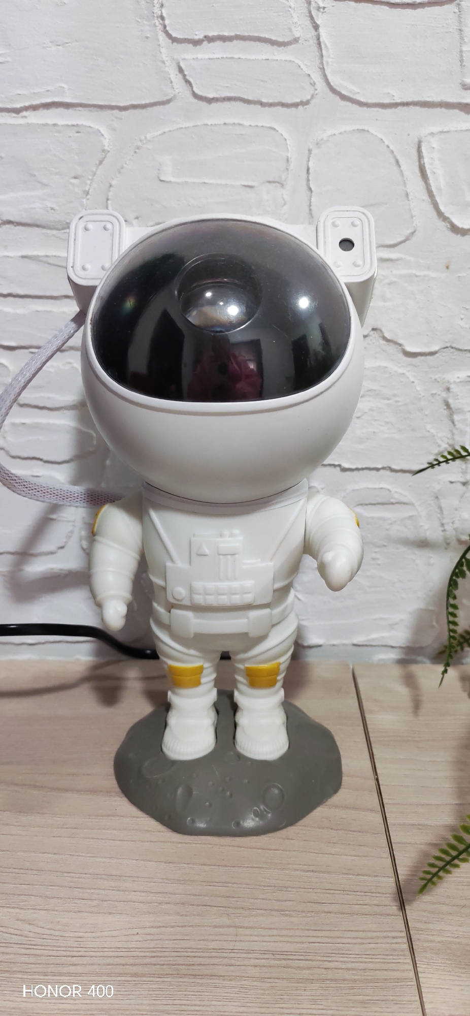 A close-up shot of a decorative astronaut figure. The figure is primarily white with yellow accents on the joints. It has a black, transparent dome-shaped helmet. The figure stands on a gray, moon-textured base. The background consists of a white brick wall and a wooden surface. A grey cable is visible to the left of the figure and a plant is partially visible to the right. The camera watermark "HONOR 400" is visible in the lower left corner. There is no indication of a location in Noviy, Russia.