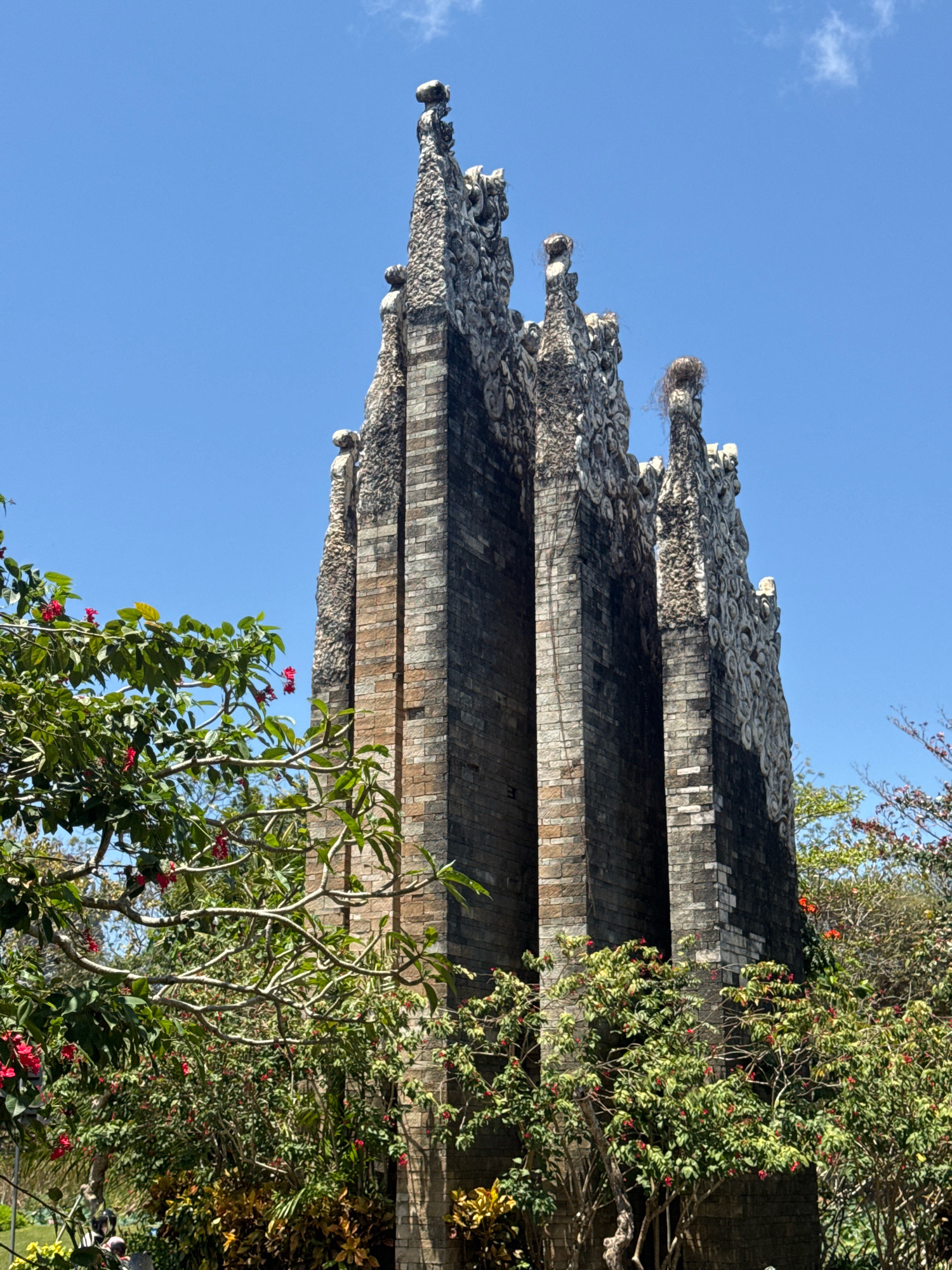 The image shows a tall, ornate stone structure in Kuta Selatan, Indonesia, against a clear blue sky. The structure appears to be made of dark brick with lighter, decorative stone accents on its upper portions, featuring intricate patterns and carved figures. The top of the structure is jagged and uneven, with some elements resembling nests. Lush green foliage with red flowers partially obscures the base of the structure. The scene is bathed in sunlight, indicating an outdoor setting during the day. There are no people clearly visible in the image.