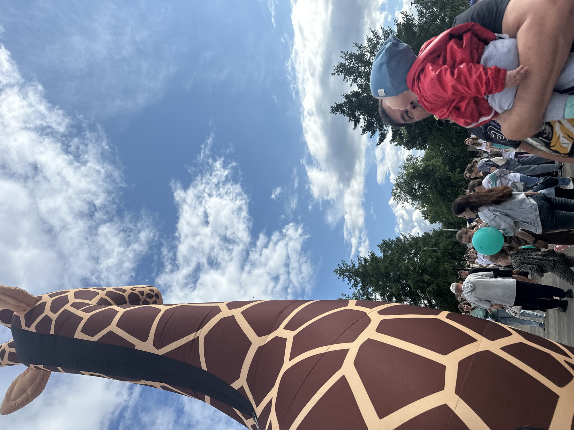 An eye-level, low-angle shot shows a giant inflatable giraffe neck dominating the left side of the frame, its brown and tan pattern stark against a bright blue sky with scattered white clouds. On the right, a man holds a baby, who is wearing a red jacket and a blue hat. The man's face is partially obscured as he leans in towards the baby. In the background, a crowd of people, some holding balloons, are gathered, and lush green trees are visible. The scene appears to be outdoors, possibly at an event or festival.